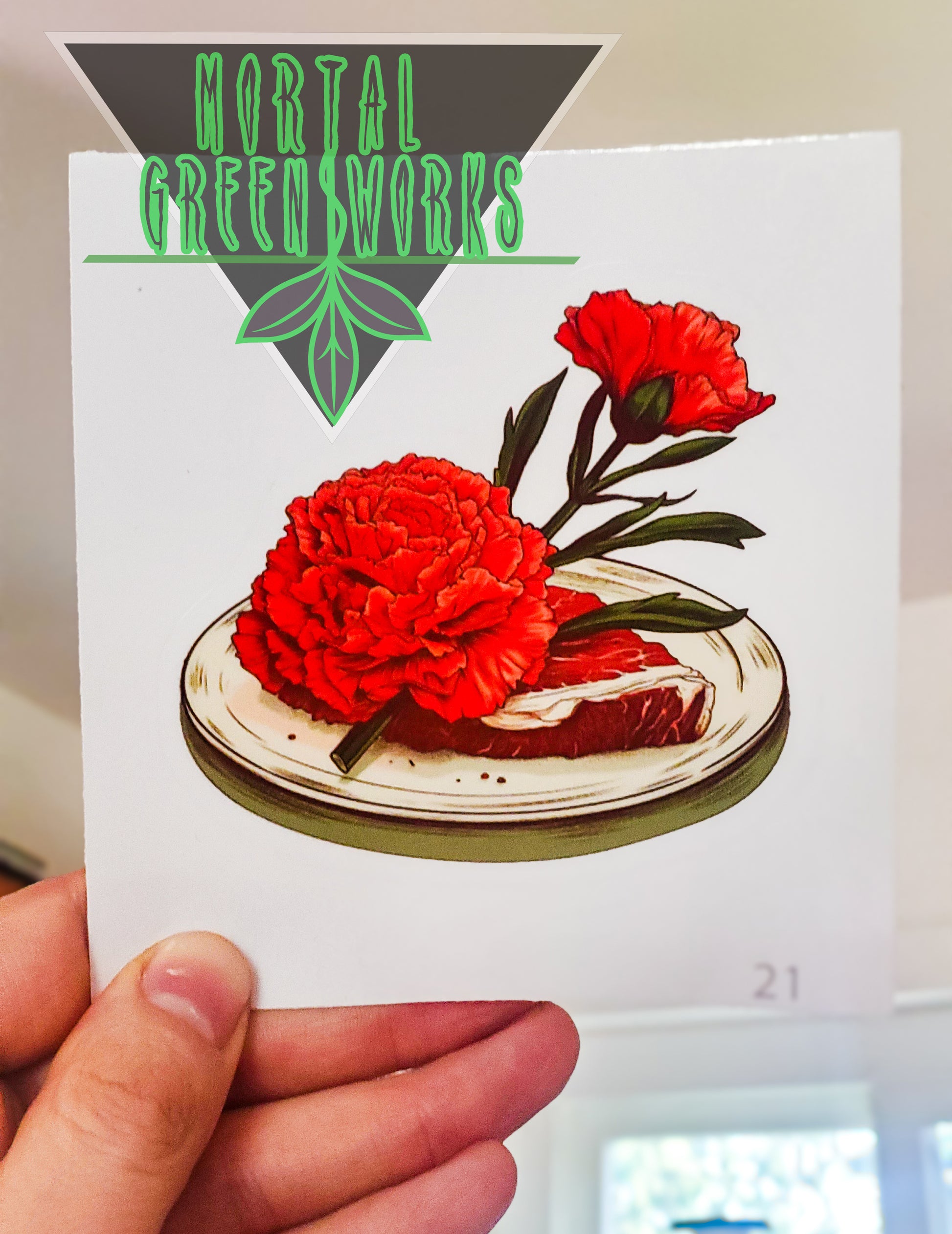 A sticker of a red carnation resting atop a raw steak, held up against an indoor background with 'Mortal Greenworks' logo.