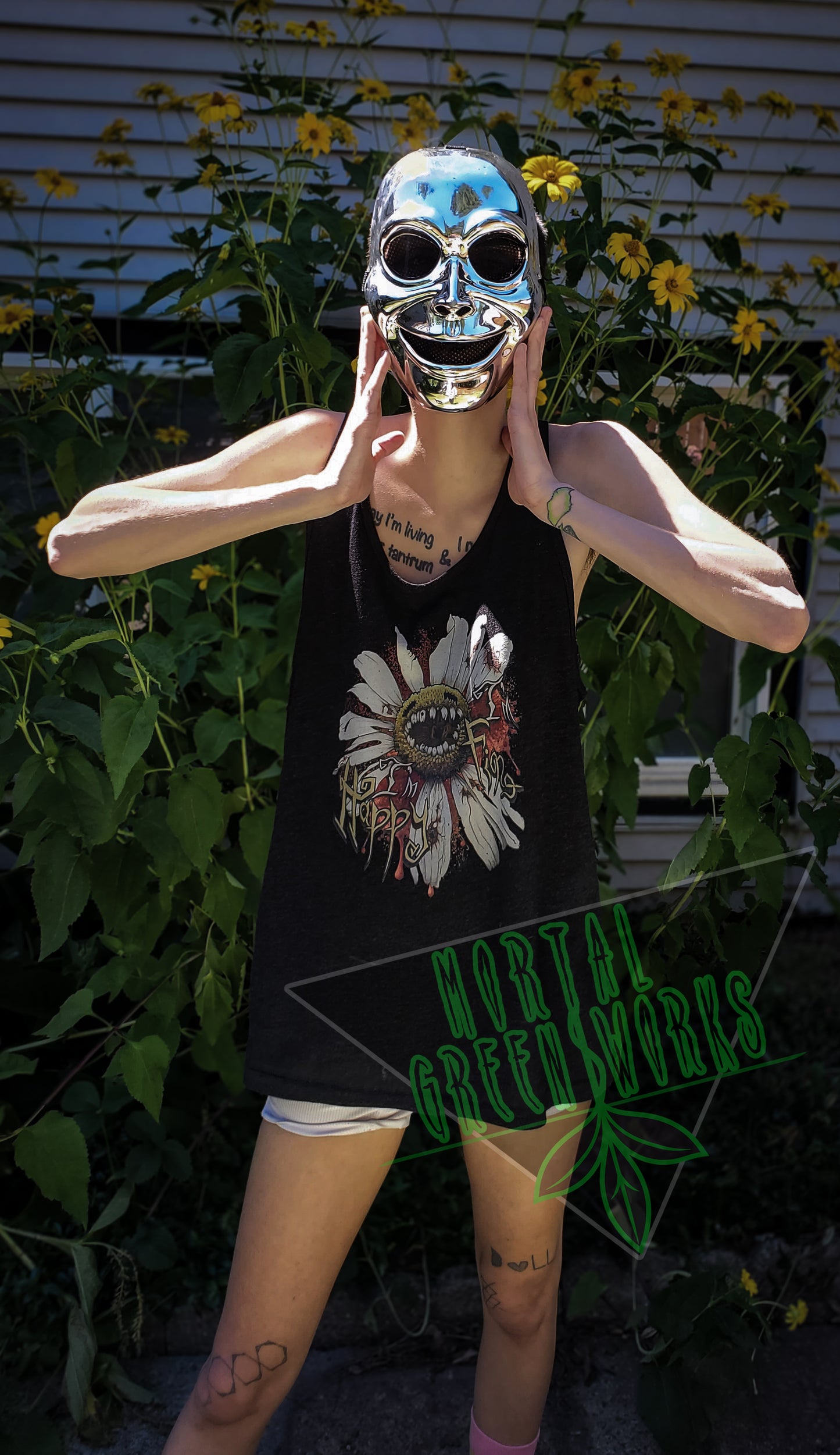 Person wearing a chrome mask and a black tank top with a "Diseased Daisy" graphic and the text "I'm Happy I'm Fine." They pose in front of tall yellow flowers. Mortal Greenworks logo overlays the image.