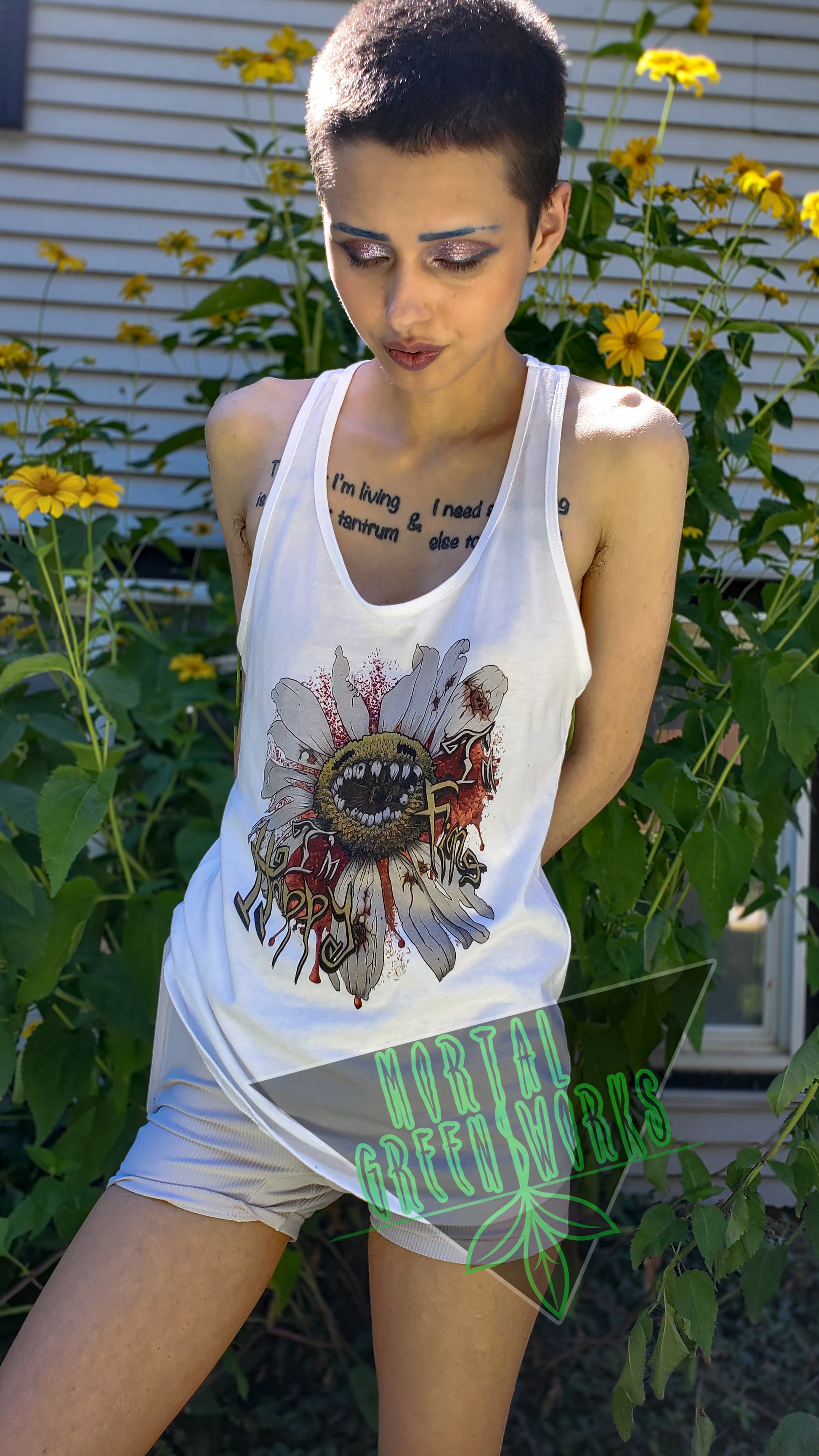 Person with short hair wearing a white tank top featuring a "Diseased Daisy" graphic with the text "I'm Happy I'm Fine." They stand in front of a garden with yellow flowers. Mortal Greenworks logo overlays the image.