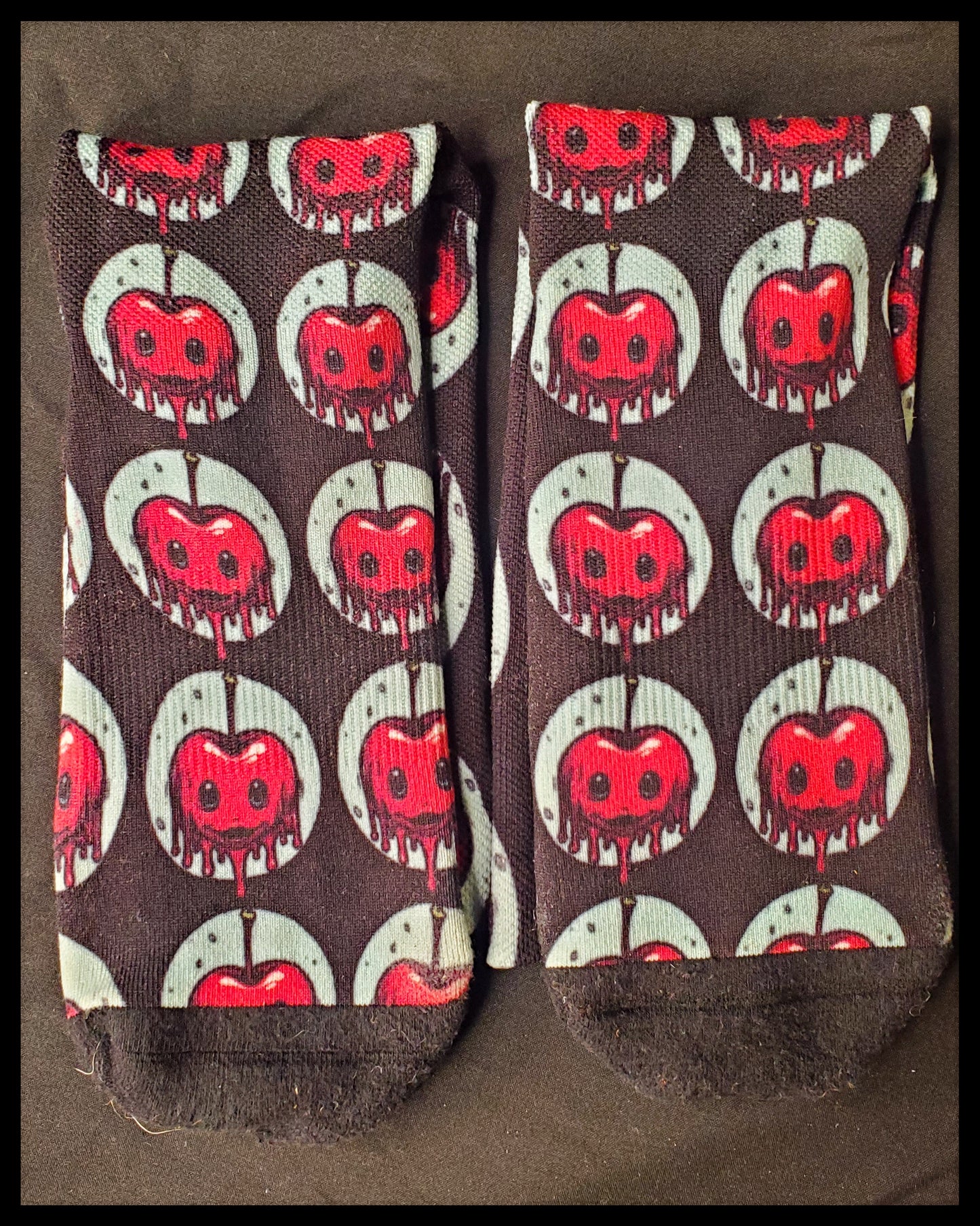 A closeup of two black socks with a red, dripping cherry with black holes for eyes and small smiles. Enclosed by a light blue circle and repeated all over the socks with the exception of the toes.