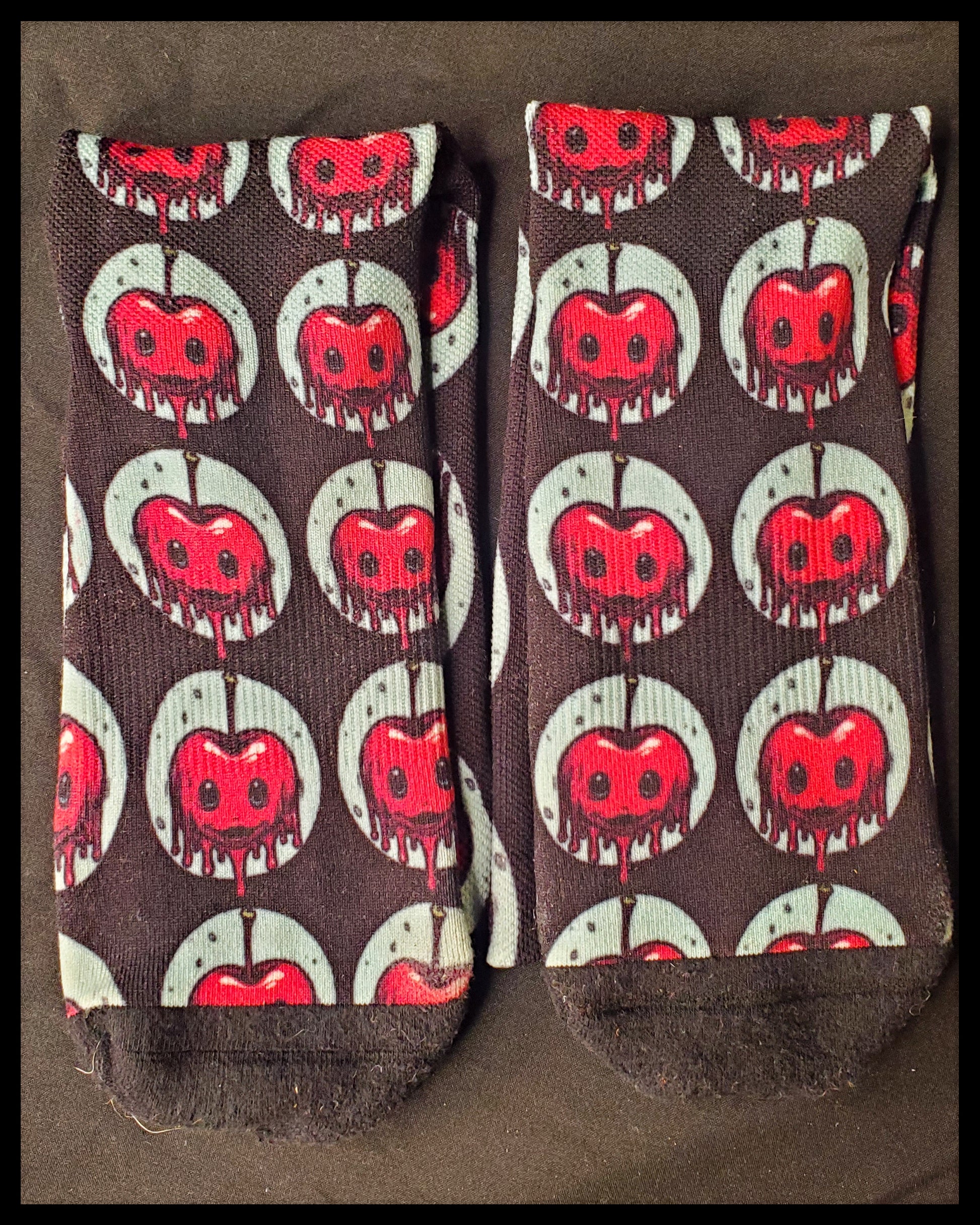 A closeup of two black socks with a red, dripping cherry with black holes for eyes and small smiles. Enclosed by a light blue circle and repeated all over the socks with the exception of the toes.