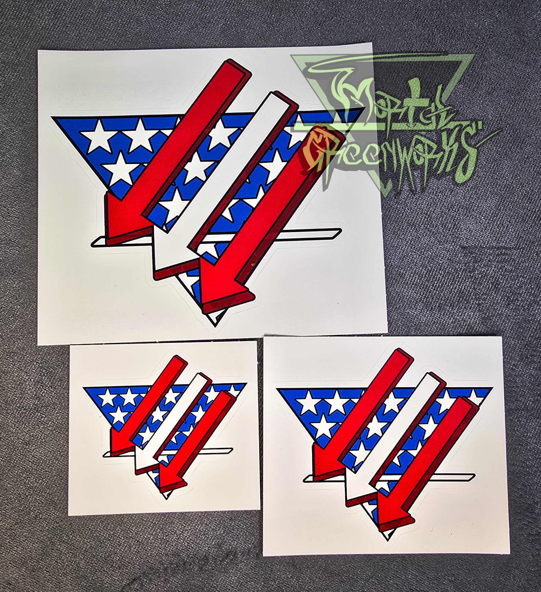 Sitting on a grey tablecloth are three stickers from big to small. The sticker design resembles an American flag is restyled as three red and white arrows pointing left and down over a triangular white and blue American flag backdrop with a bold white line in the bottom third. The Mortal Greenworks Logo is displayed in the top right of the photo.