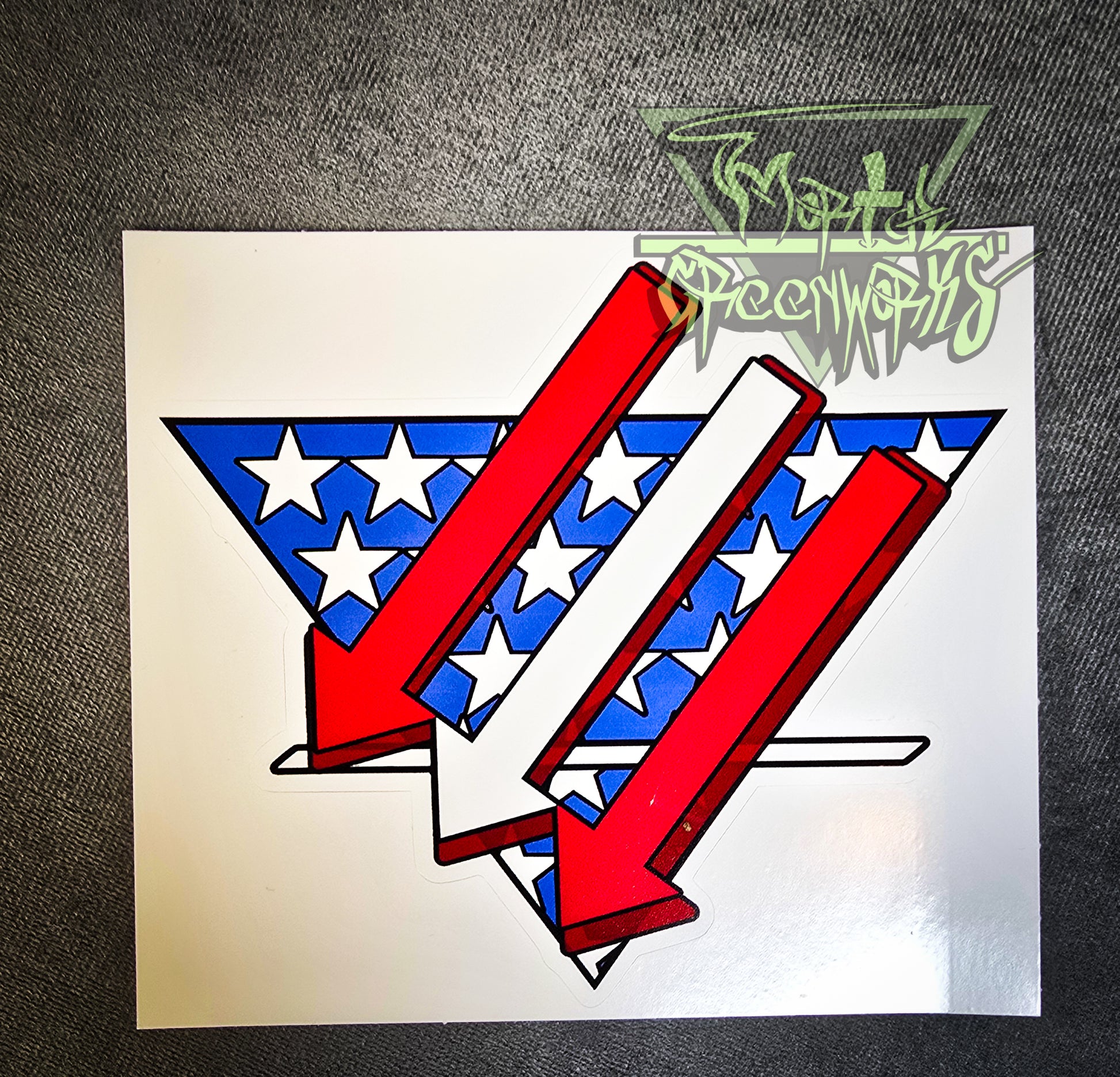 Sitting on a grey tablecloth is a  6x6" sticker resembling an American flag is restyled as three red and white arrows pointing left and down over a triangular white and blue American flag backdrop with a bold white line in the bottom third. The Mortal Greenworks Logo is displayed in the top right of the photo.