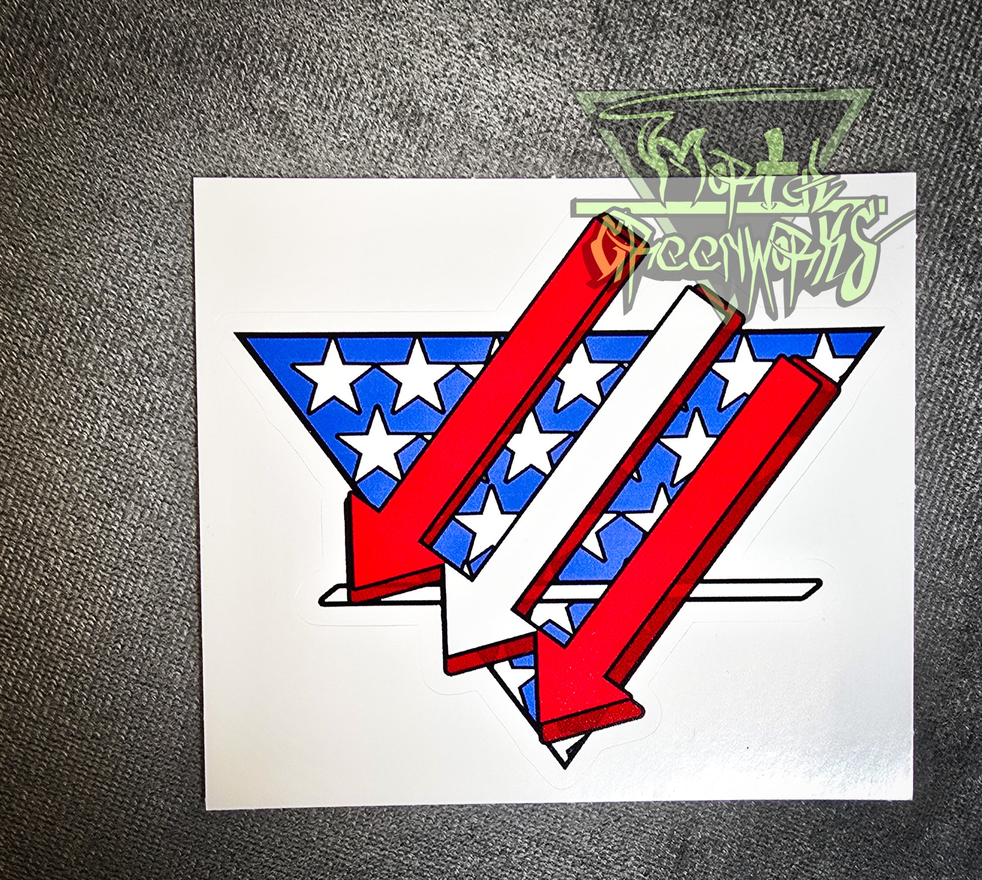 Sitting on a grey tablecloth is a  3x3" sticker resembling an American flag is restyled as three red and white arrows pointing left and down over a triangular white and blue American flag backdrop with a bold white line in the bottom third. The Mortal Greenworks Logo is displayed in the top right of the photo.