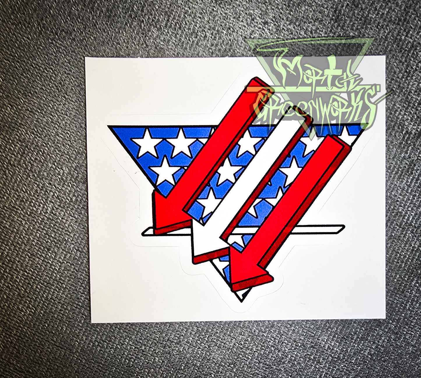 Sitting on a grey tablecloth is a  3x3" sticker resembling an American flag is restyled as three red and white arrows pointing left and down over a triangular white and blue American flag backdrop with a bold white line in the bottom third. The Mortal Greenworks Logo is displayed in the top right of the photo.