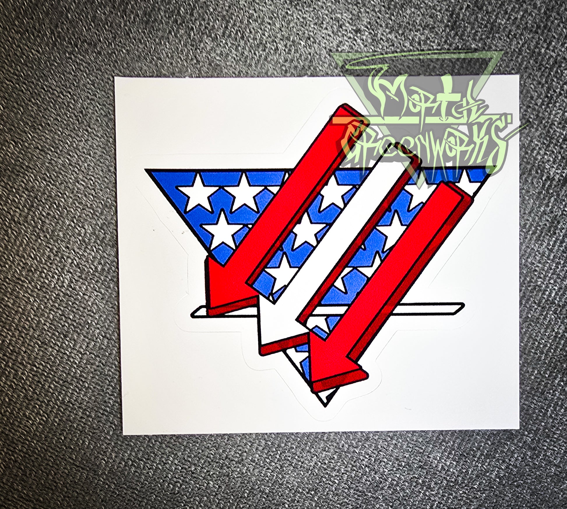 Sitting on a grey tablecloth is a  3x3" sticker resembling an American flag is restyled as three red and white arrows pointing left and down over a triangular white and blue American flag backdrop with a bold white line in the bottom third. The Mortal Greenworks Logo is displayed in the top right of the photo.