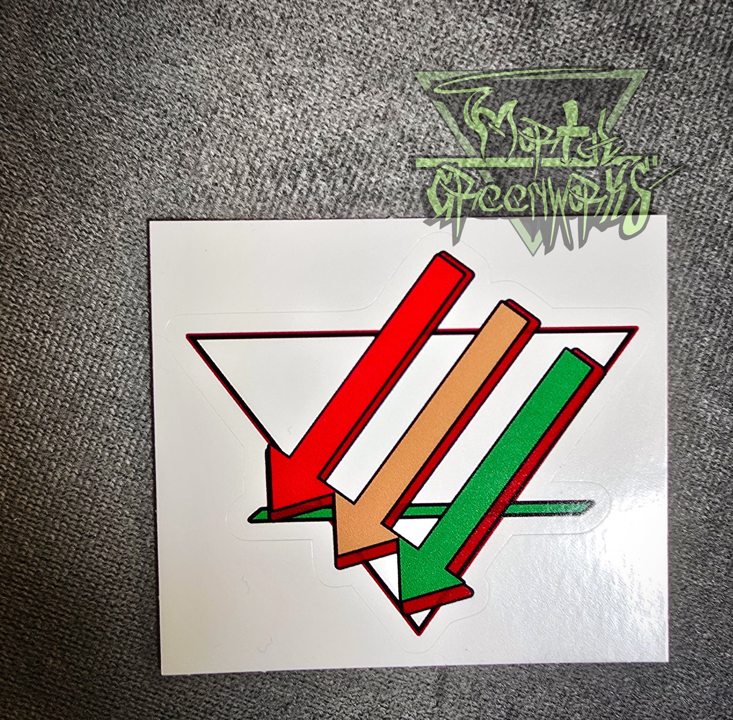 A close-up of a 3x3" sticker sitting on a dark tablecloth containing three arrows pointing downward and left, colored red, brown, and green. Overlapping an upside-down white triangle with a bold green line in the bottom third. The Mortalgreenworks Logo sits in the top right hand corner of the photograph