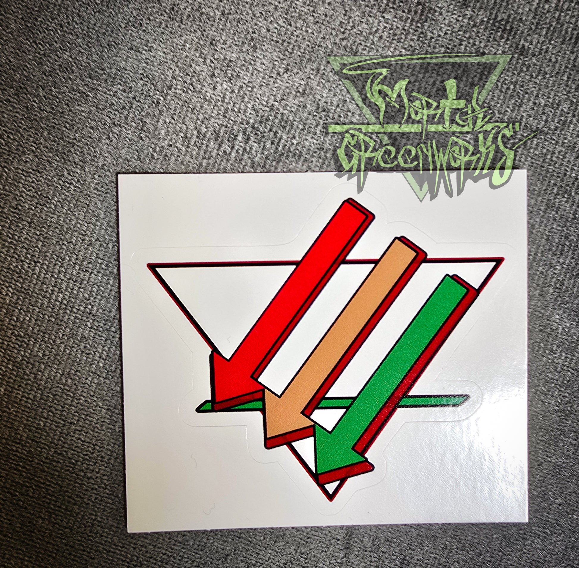 A close-up of a 3x3" sticker sitting on a dark tablecloth containing three arrows pointing downward and left, colored red, brown, and green. Overlapping an upside-down white triangle with a bold green line in the bottom third. The Mortalgreenworks Logo sits in the top right hand corner of the photograph