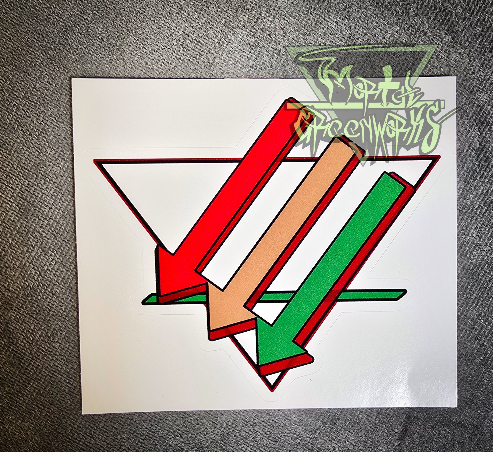 A close-up of a 4x4 sticker sitting on a dark tablecloth containing three arrows pointing downward and left, colored red, brown, and green. Overlapping an upside-down white triangle with a bold green line in the bottom third. The Mortalgreenworks Logo sits in the top right-hand corner of the photograph