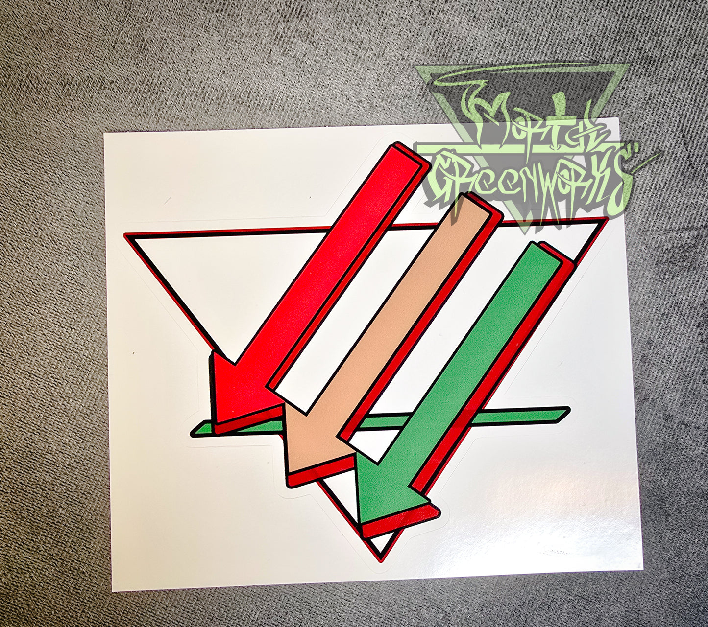 A close-up of a 6x6 sticker sitting on a dark tablecloth containing three arrows pointing downward and left, colored red, brown, and green. Overlapping an upside-down white triangle with a bold green line in the bottom third. The Mortalgreenworks Logo sits in the top right hand corner of the photograph