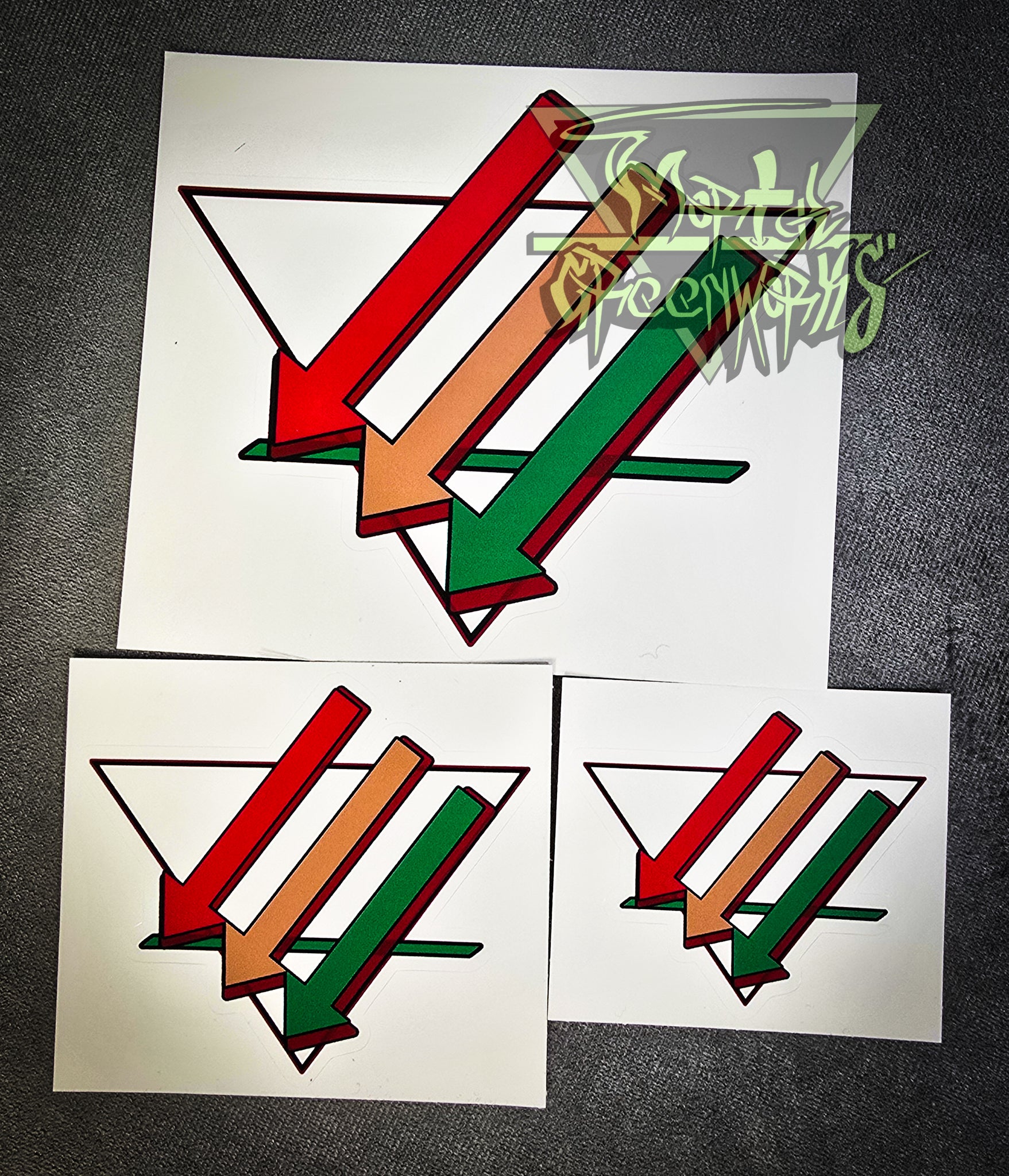 A close up photograph of three stickers on a dark tablecloth. The sticker graphic is three arrows pointing to downward and left colored red, brown, and green. Overlapping an upside down white triangle with a bold green line in the bottom third. The sticker sizes go from big to small.