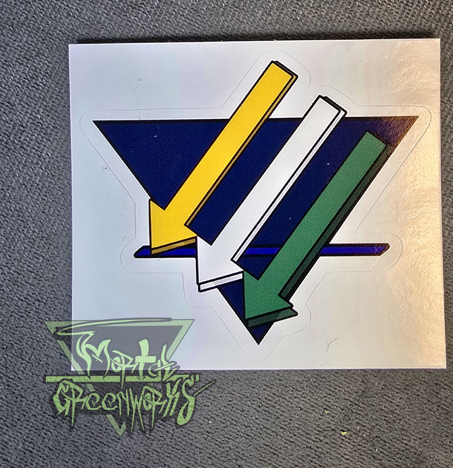 Close up of a 3x3" sticker showing the three iron cross down arrows painted gold, white, nd green, are on aadark blue upside down triangle with a lighter blue bold line in the bottom third T.he Mortal Greenworks Logo sits in the bottom left corner
