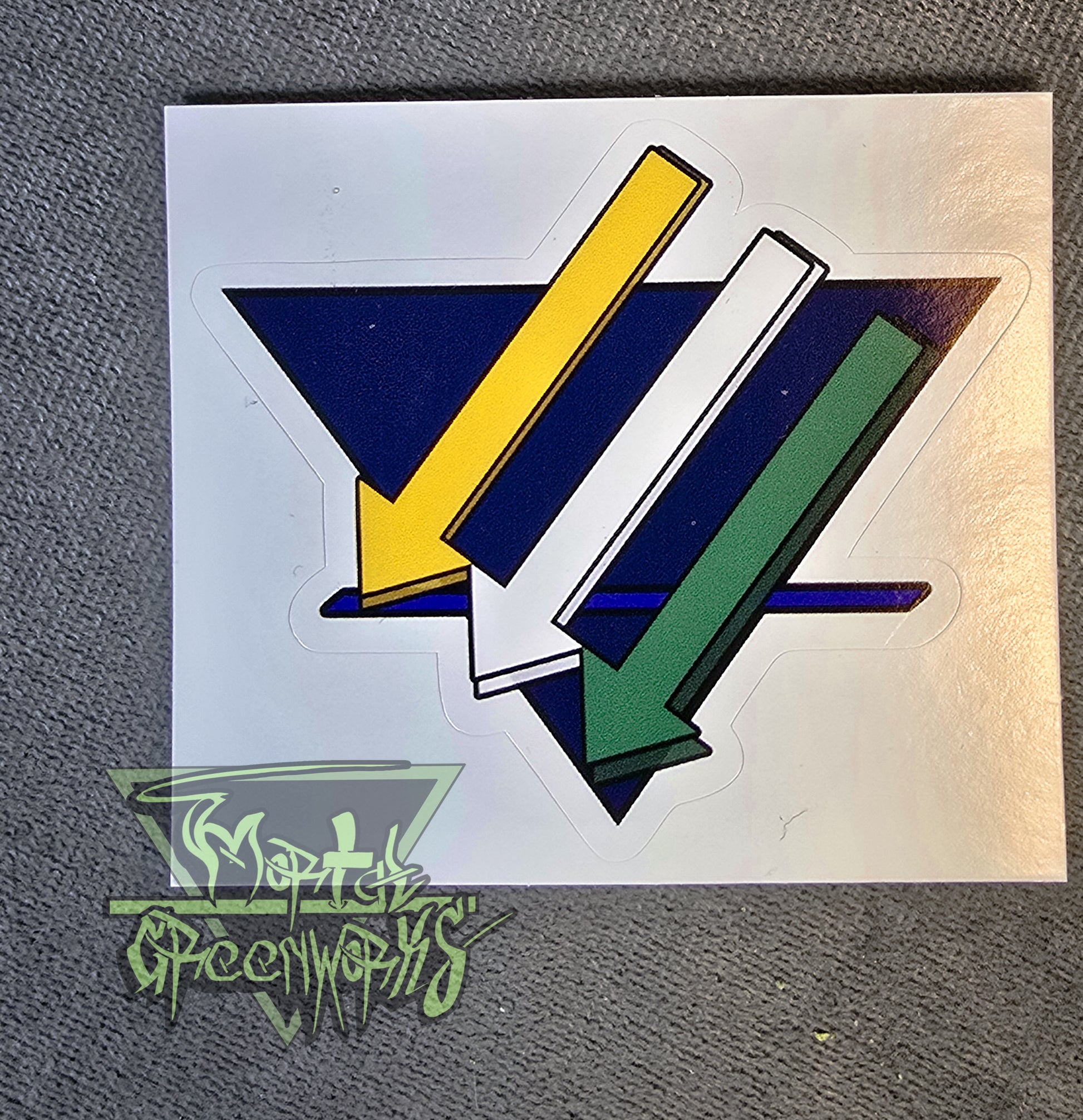 Close up of a 3x3" sticker showing the three iron cross down arrows painted gold, white, nd green, are on aadark blue upside down triangle with a lighter blue bold line in the bottom third T.he Mortal Greenworks Logo sits in the bottom left corner