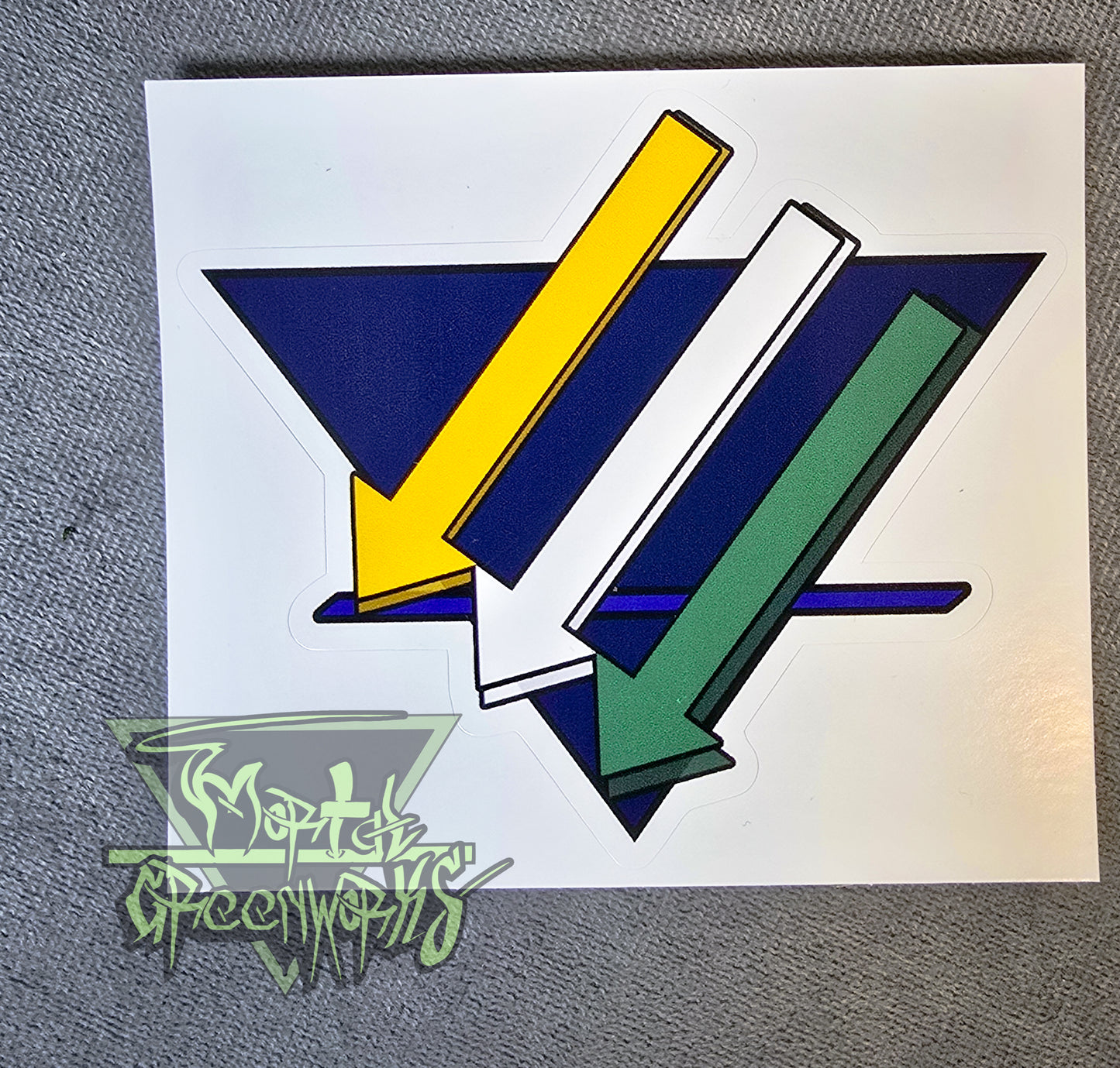 Close up of a 4x4" sticker showing the three iron cross down arrows painted gold, white a,nd green, are on aadark blue upside down triangle with a lighter blue bold line in the bottom third T.he Mortal Greenworks Logo sits in the bottom left corner
