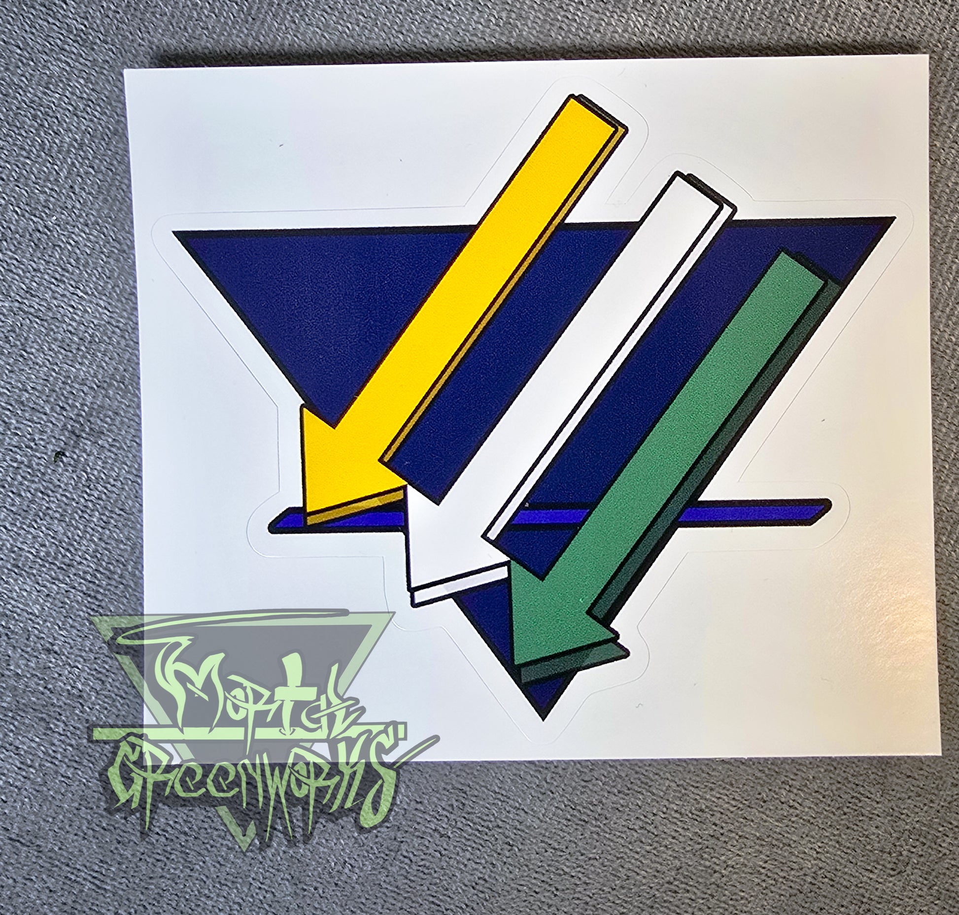 Close up of a 4x4" sticker showing the three iron cross down arrows painted gold, white a,nd green, are on aadark blue upside down triangle with a lighter blue bold line in the bottom third T.he Mortal Greenworks Logo sits in the bottom left corner