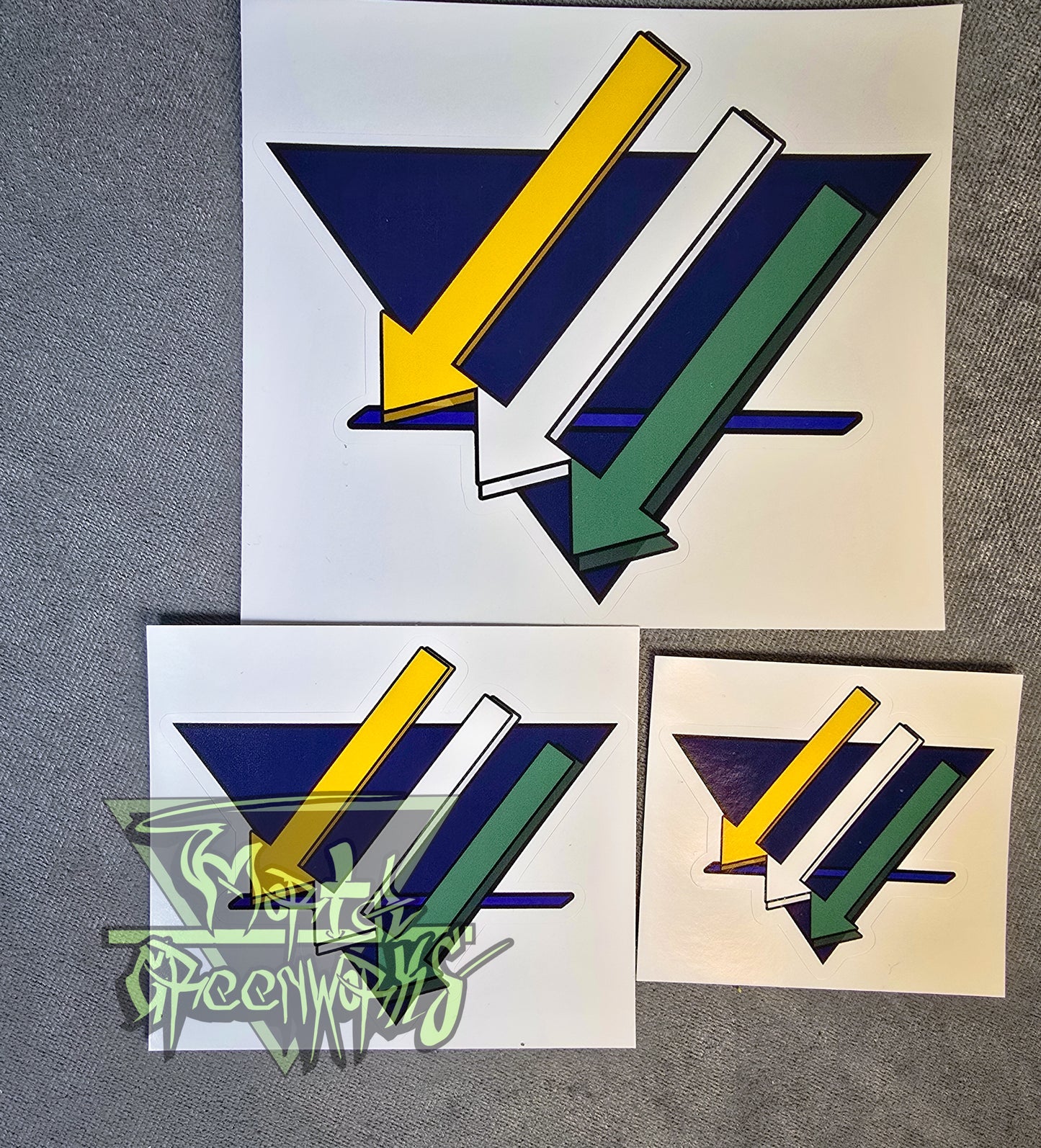 Three stickers of the gold, white, and green downward arrows on the blue triangle rest on a grey table cloth. Pictured side by side showing 3x3",4x4" and 6x"6 as well as the transparent Mortal Greenworks logo in the bottom left corner of the photo.