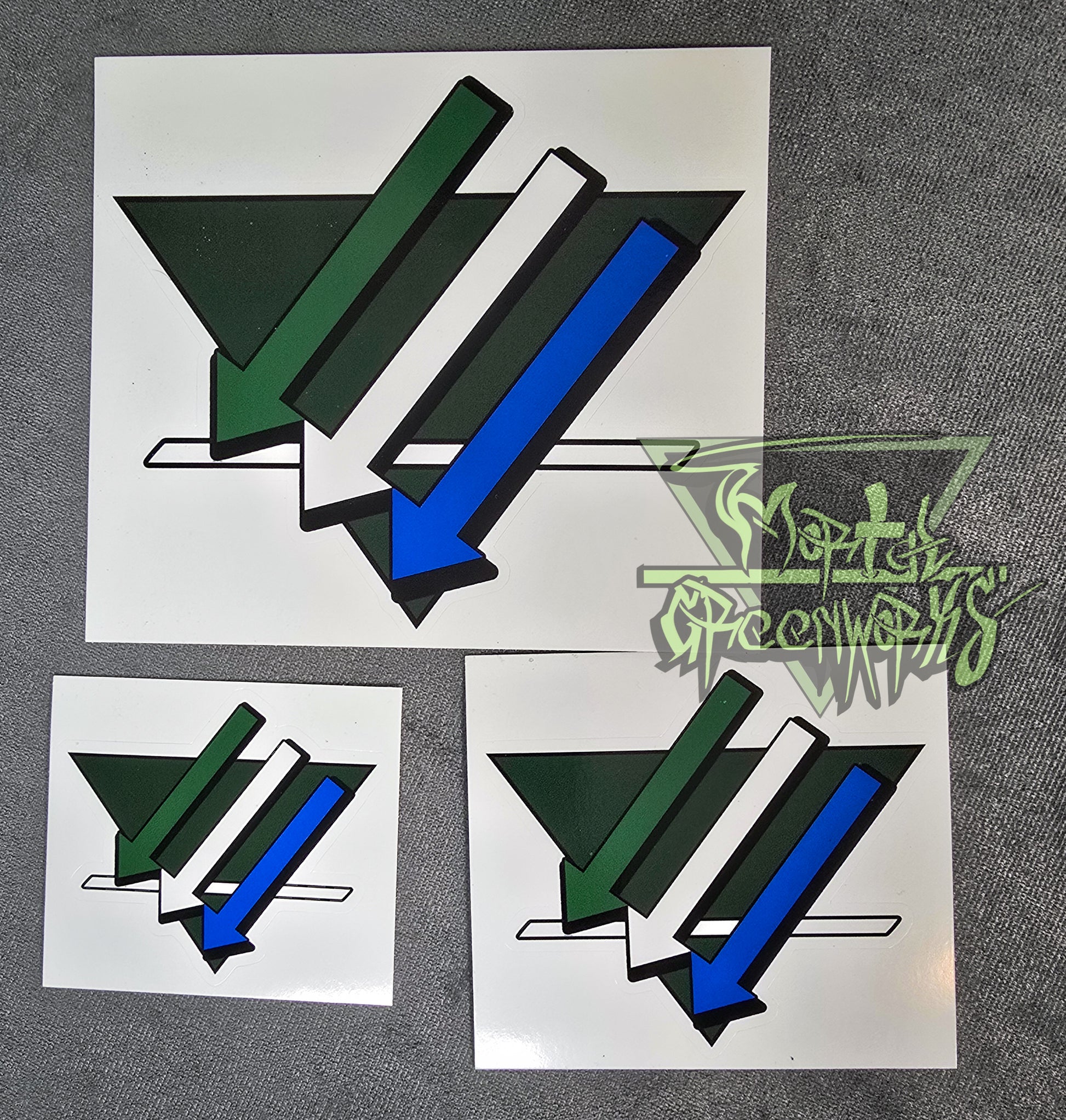 Photograph of a set of three stickers of different sizes. The same forest-green triangular sticker with three arrows and a white line overlay. The arrows are green, white, and blue from top to bottom. corner.