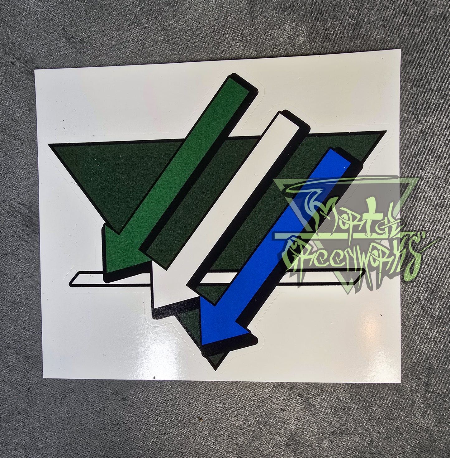 Photograph of a forest green 4x4" triangular sticker with three arrows and a white line overlaying. The arrows are from top to bottom green, white and blue.