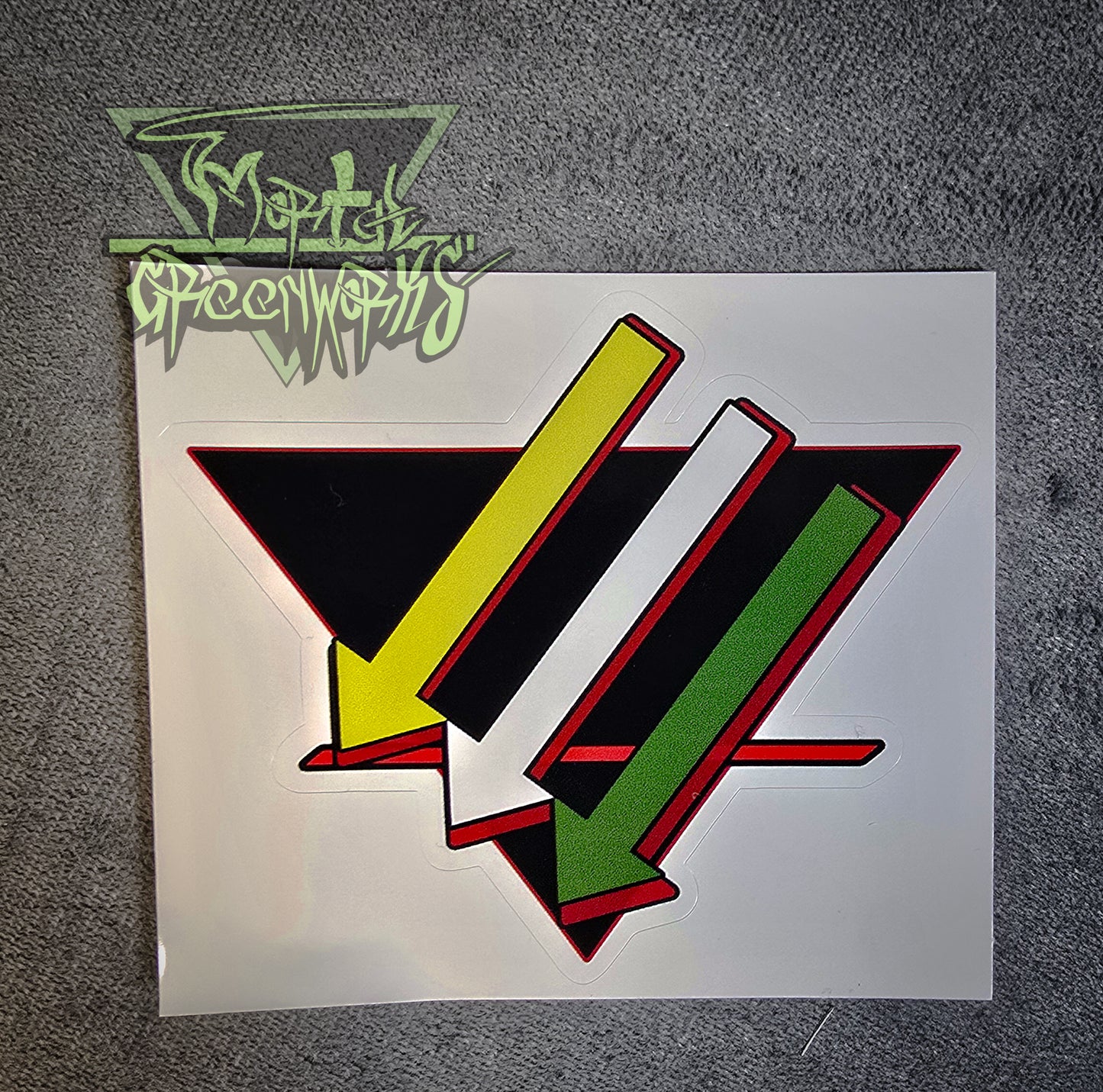 A dim close-up photograph of a 4x4 sticker on a grey tablecloth. Three arrows point down and to the left, parallel to each other, the first one gold, the second silver, and the third green. They lay over a black, upside-down triangle with a bold red line in the bottom third. The sticker has a red outline. The Mortal Greenworks watermark is in the top-left corner of the image.