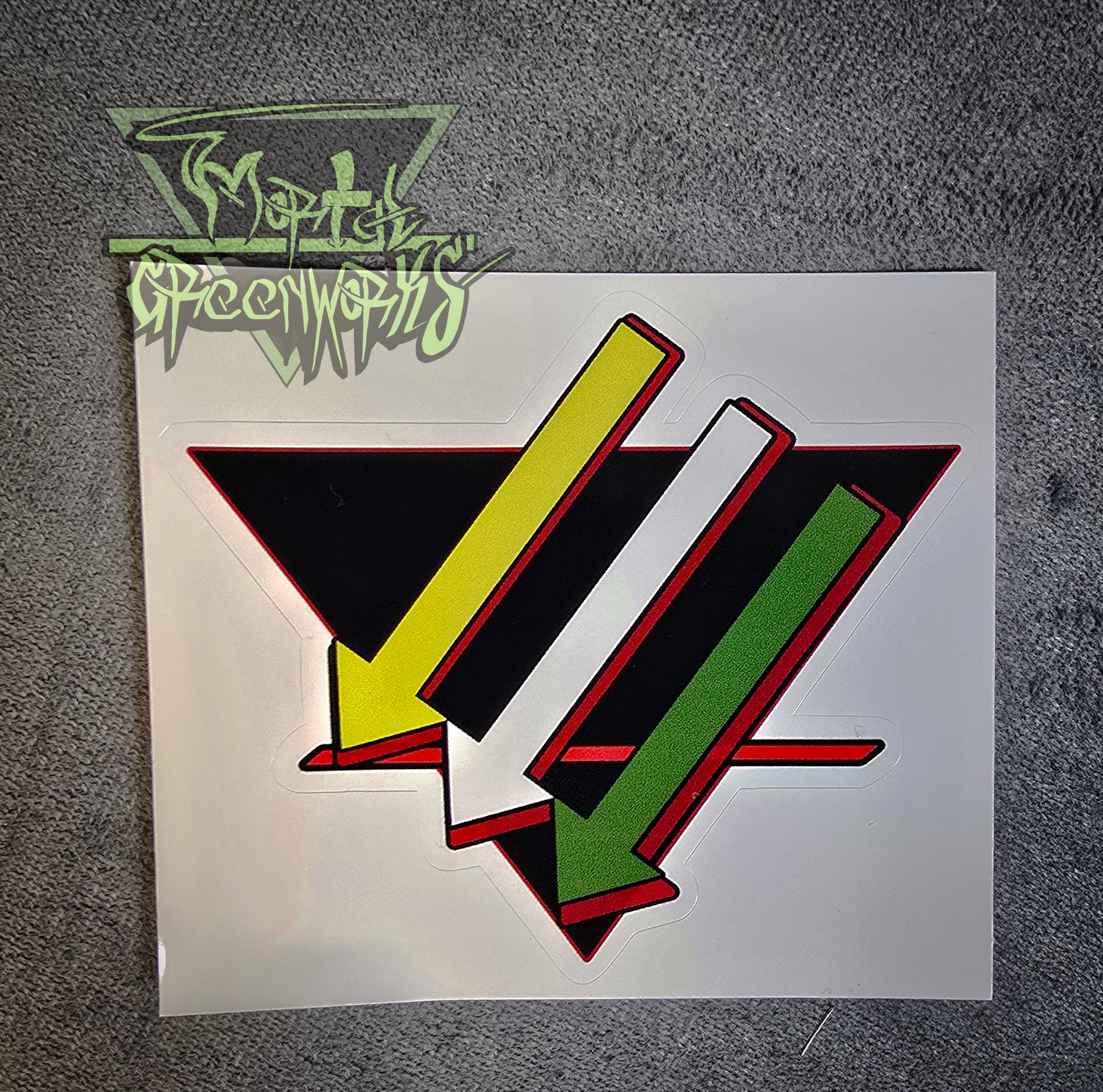 A dim close-up photograph of a 4x4 sticker on a grey tablecloth. Three arrows point down and to the left, parallel to each other, the first one gold, the second silver, and the third green. They lay over a black, upside-down triangle with a bold red line in the bottom third. The sticker has a red outline. The Mortal Greenworks watermark is in the top-left corner of the image.