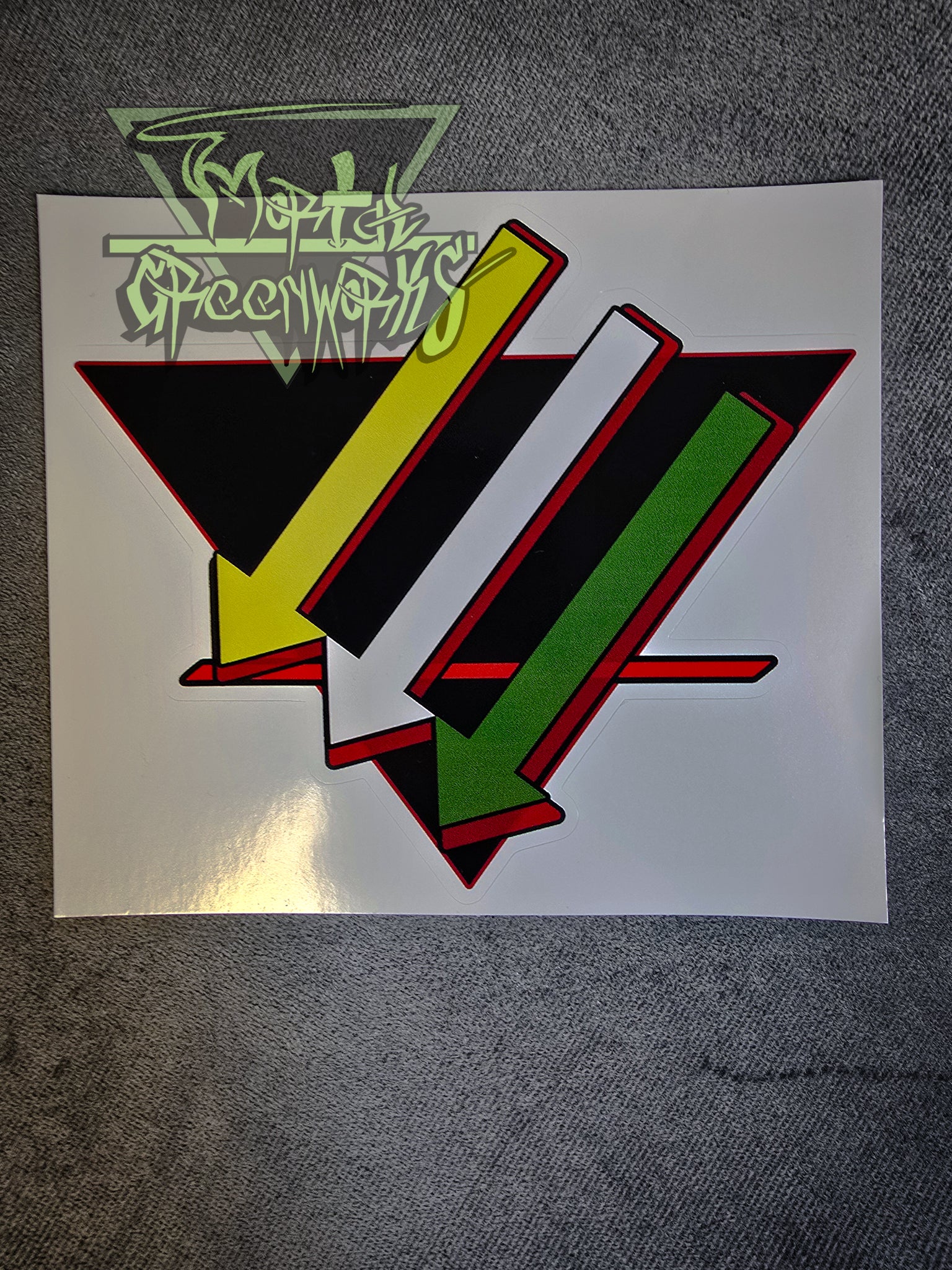 A dim close-up photograph of a 6x6 sticker on a grey tablecloth. Three arrows point down and to the left, parallel to each other, the first one gold, the second silver, and the third green. They lay over a black, upside-down triangle with a bold red line in the bottom third. The sticker has a red outline. The Mortal Greenworks watermark is in the top-left corner of the image.