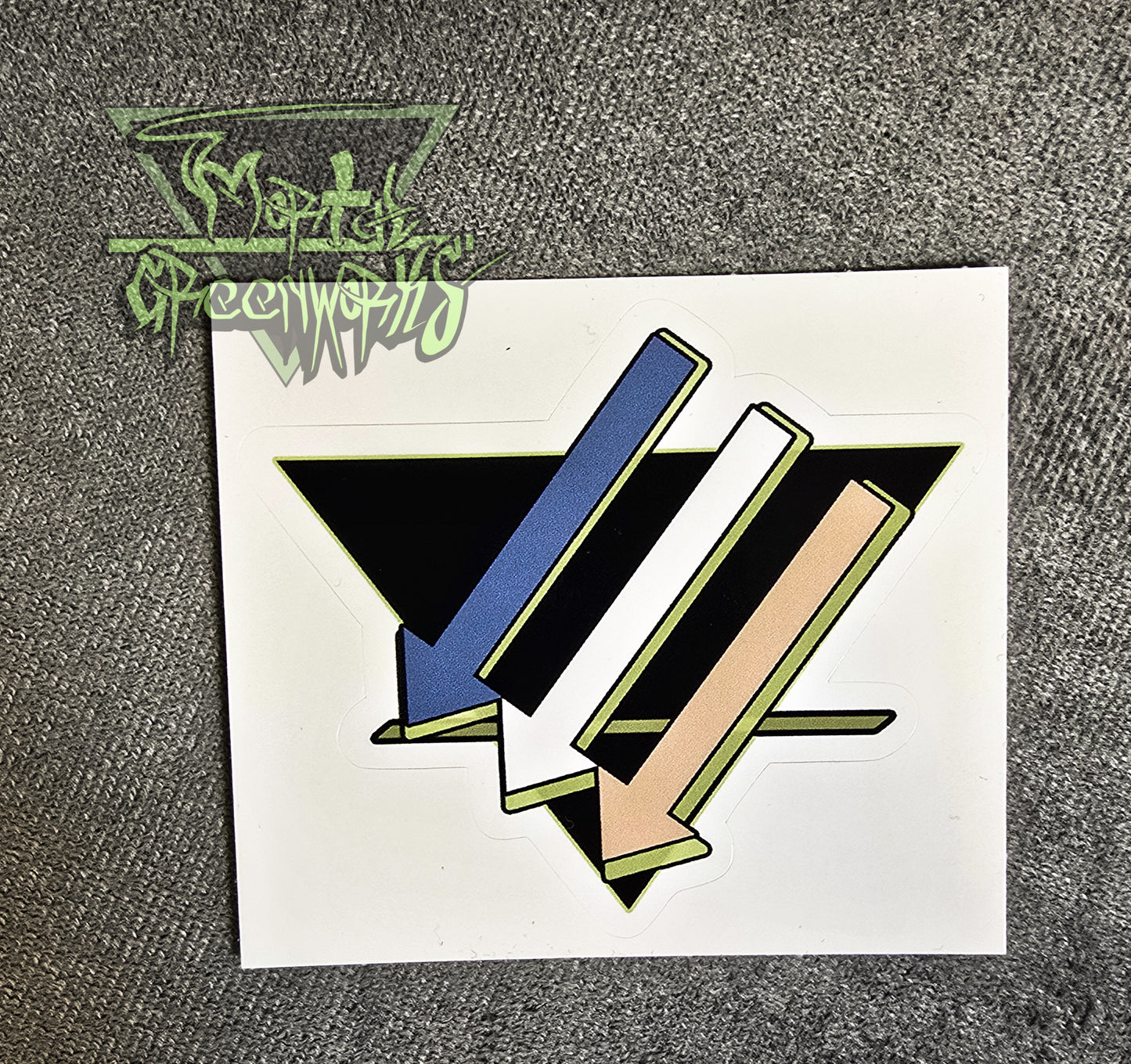 A 3x3 sticker of three downward-pointing arrows pointing left is colored light blue, white, and tan, with a light green highlight, and an upside-down black triangle with a green line on the bottom third rests on a grey tablecloth. The Mortal greenworks logo sits in the top-left corner of the photo.