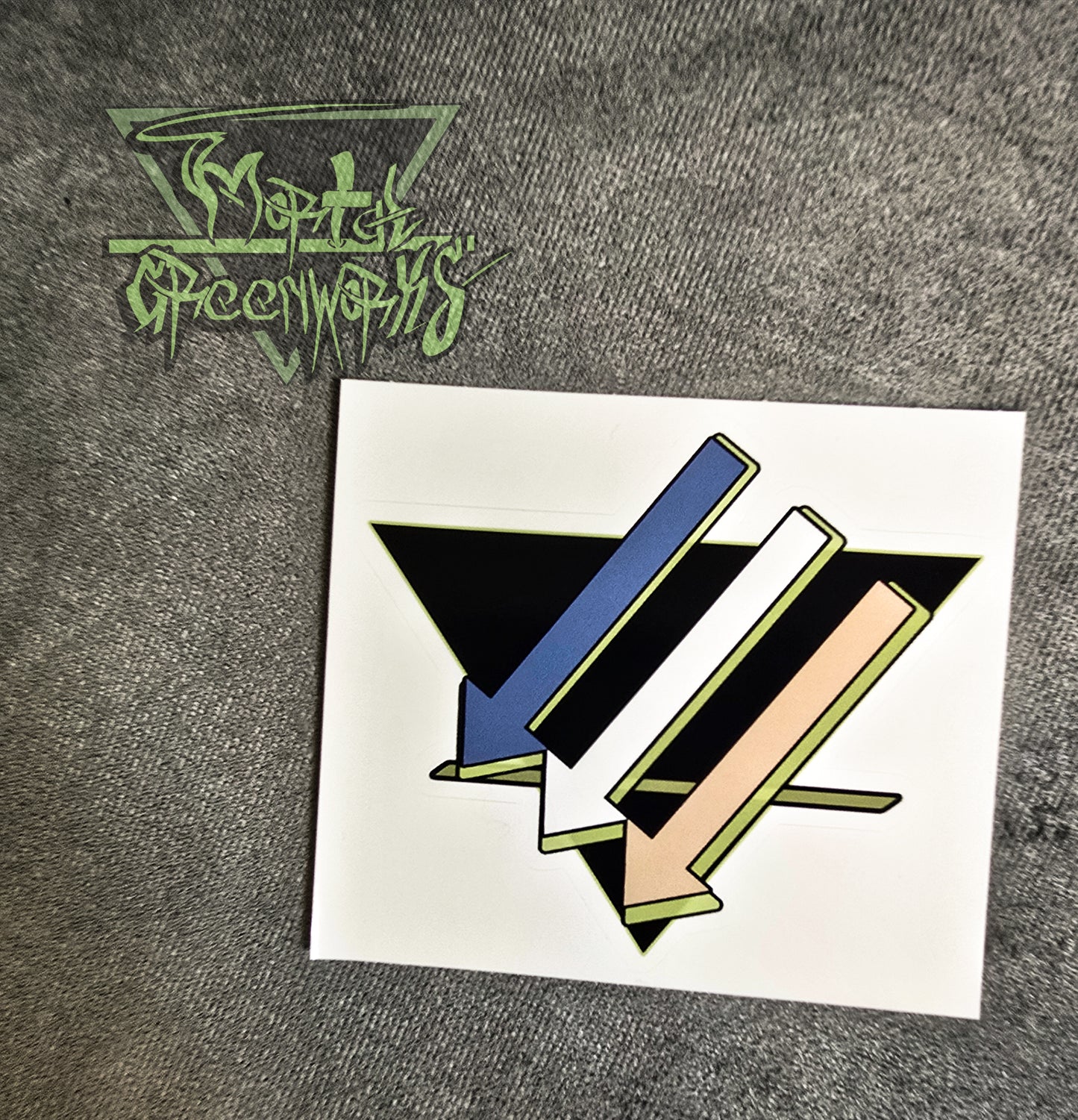 A 4x4 sticker of three downward-pointing arrows pointing left is colored light blue, white, and tan, with a light green highlight, and an upside-down black triangle with a green line on the bottom third rests on a grey tablecloth. The Mortal greenworks logo sits in the top-left corner of the photo.
