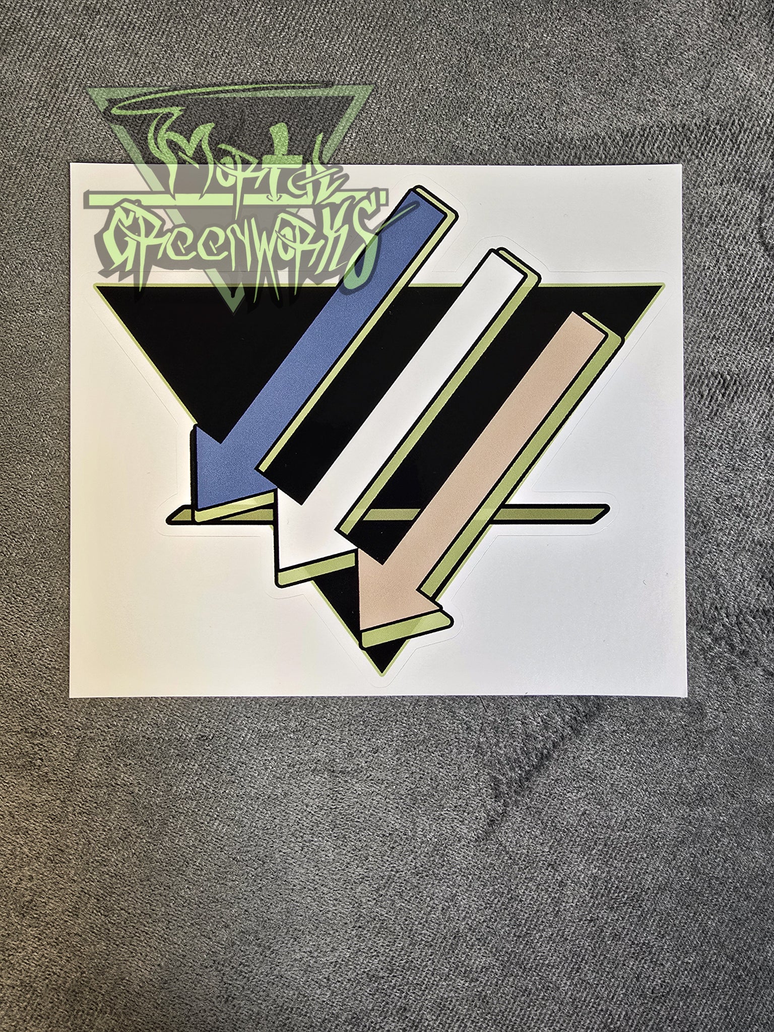 A 6x6 sticker of three downward-pointing arrows pointing left is colored light blue, white, and tan, with a light green highlight, and an upside-down black triangle with a green line on the bottom third rests on a grey tablecloth. The Mortal greenworks logo sits in the top-left corner of the photo.