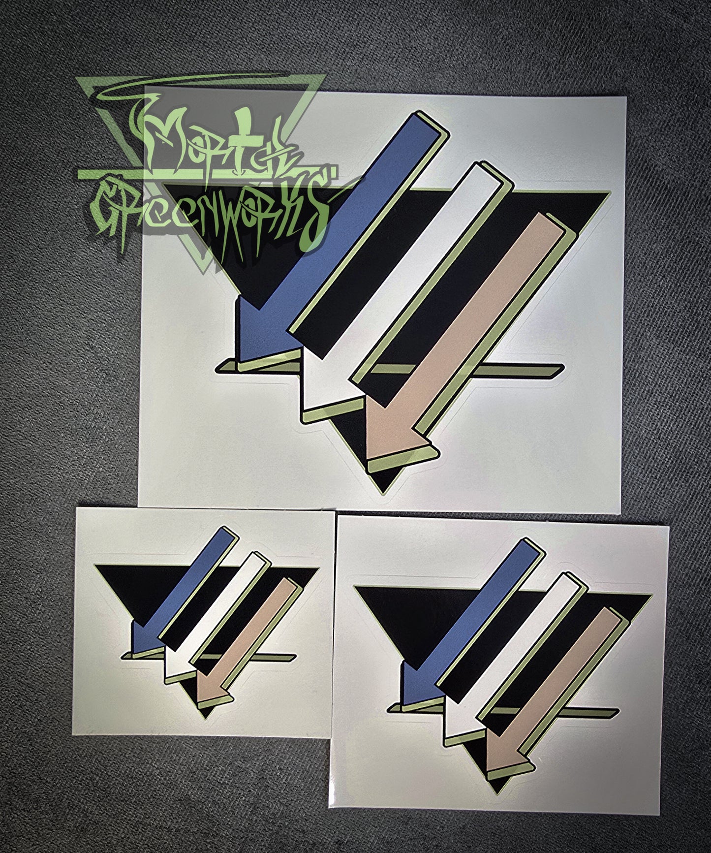Three stickers of three downward arrows pointing left are colored light blue, white, and tan, with a light green highlight, with an upside-down black triangle with a green line on the bottom third, resting on a grey tablecloth. Pictured side by side showing 3x3",4x4" and 6x"6 as well as the transparent Mortal Greenworks logo in the bottom left corner of the photo.