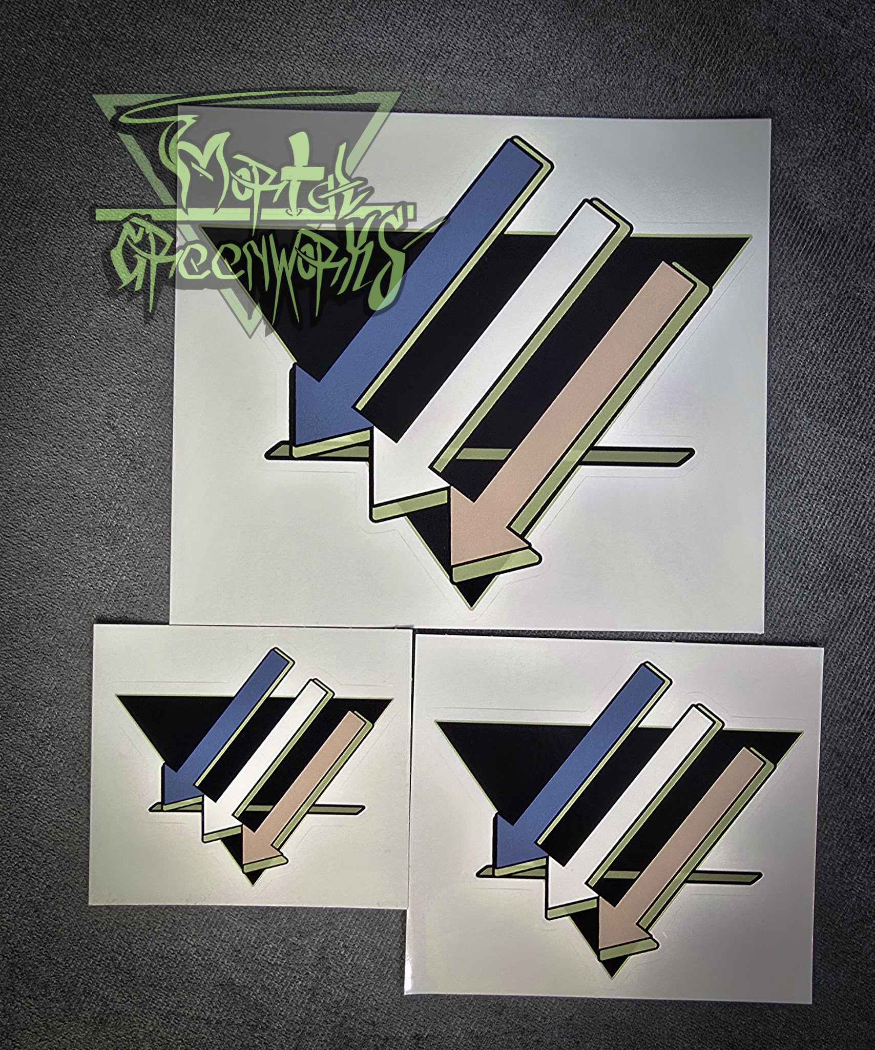 Three stickers of three downward arrows pointing left are colored light blue, white, and tan, with a light green highlight, with an upside-down black triangle with a green line on the bottom third, resting on a grey tablecloth. Pictured side by side showing 3x3",4x4" and 6x"6 as well as the transparent Mortal Greenworks logo in the bottom left corner of the photo.