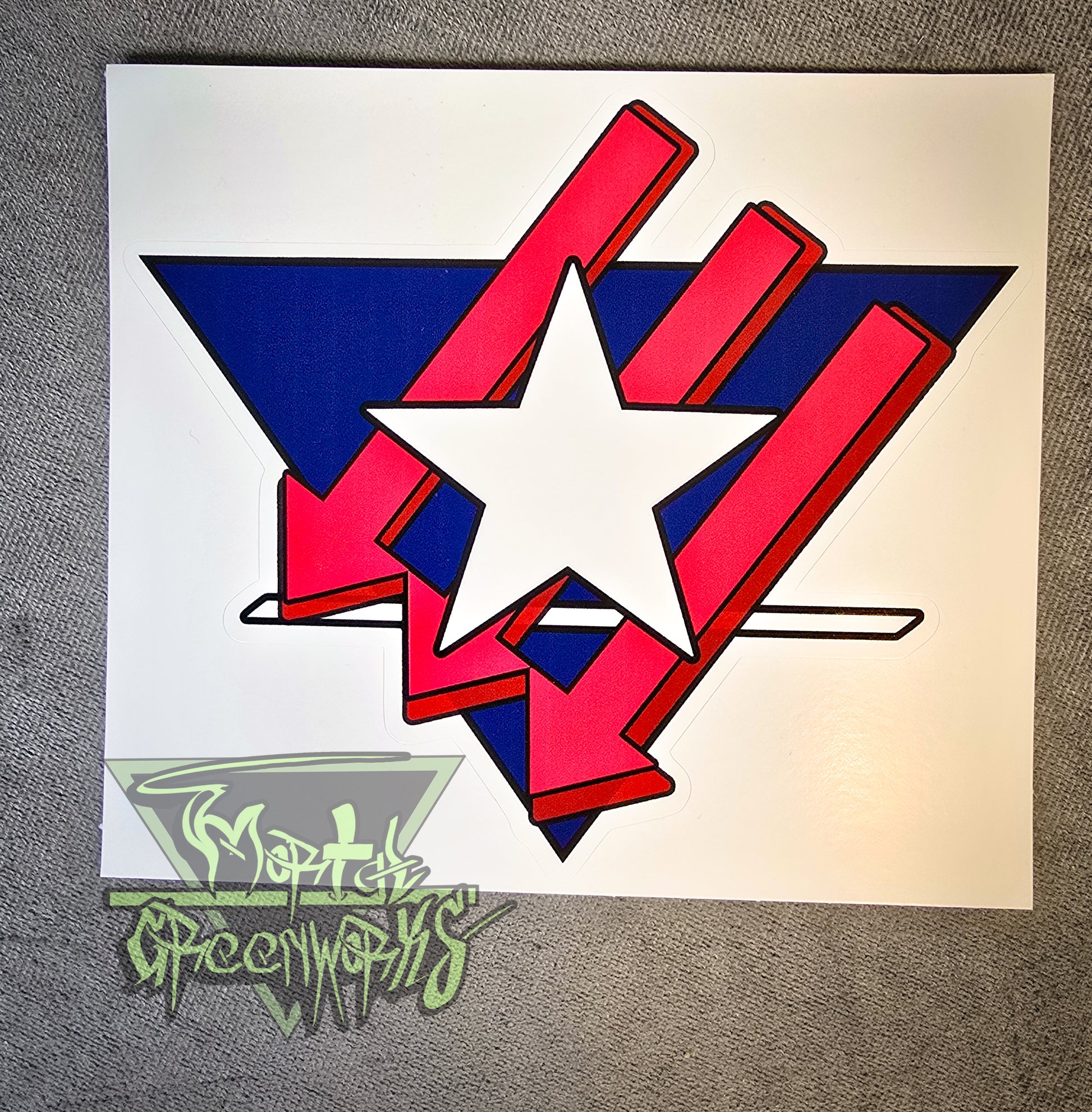 A 3x3" sticker sits on top of a grey tablecloth. It features a Texas Lone Star design atop three red iron front arrows, all enclosed in a navy blue upside-down triangle with a white line at the bottom third. The Mortal Greenworks Logo sits in the bottom left corner.