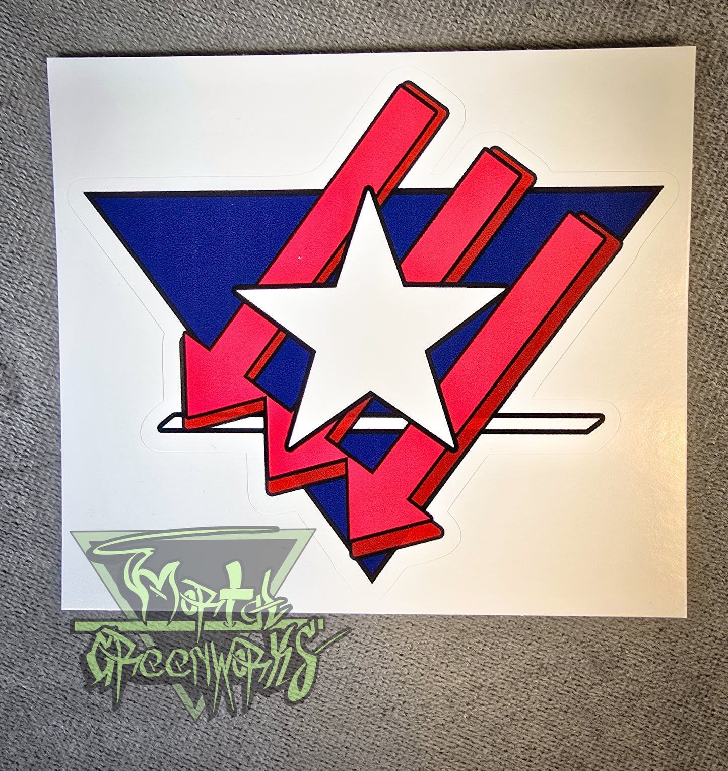 A 4x4" sticker sits on top of a grey tablecloth. It features a Texas Lone Star design atop three red iron front arrows, all enclosed in a navy blue upside-down triangle with a white line at the bottom third. The Mortal Greenworks Logo sits in the bottom left corner.
