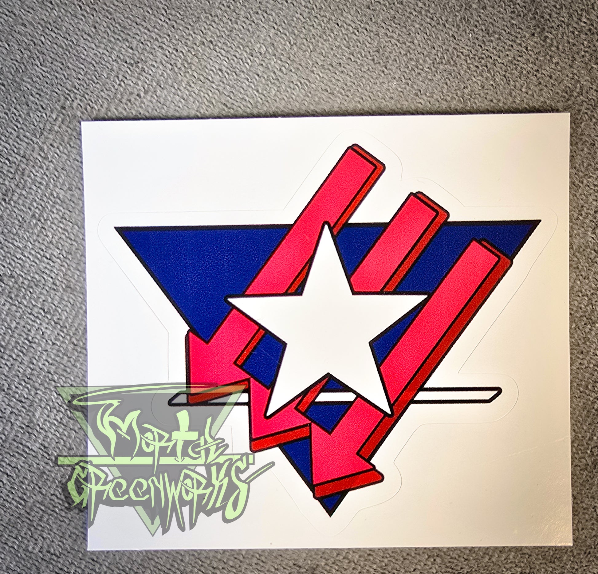 A 6x6" sticker sits on top of a grey tablecloth. It features a Texas Lone Star design atop three red iron front arrows, all enclosed in a navy blue upside-down triangle with a white line at the bottom third. The Mortal Greenworks Logo sits in the bottom left corner.