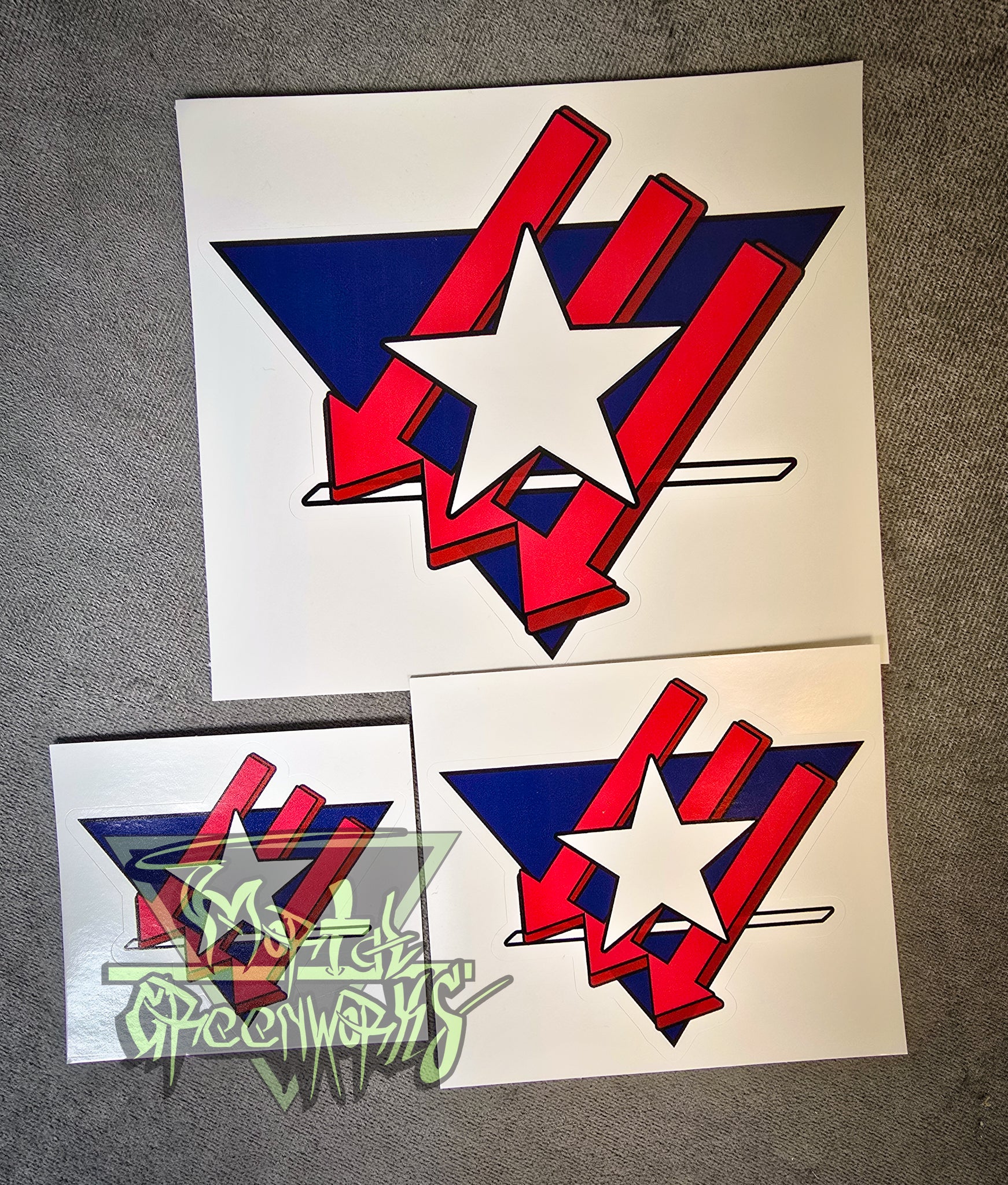Three stickers sit on a grey tablecloth, showing the white Texas Lone Sar along with the Iron Front logo painted red, cast over an upside down navy blue triangle with a bold white line in the bottom third. The Mortal Greenworks logo is in the bottom left corner. All three sizes, from 3x3", 4x4", and 6x6", are displayed side by side.