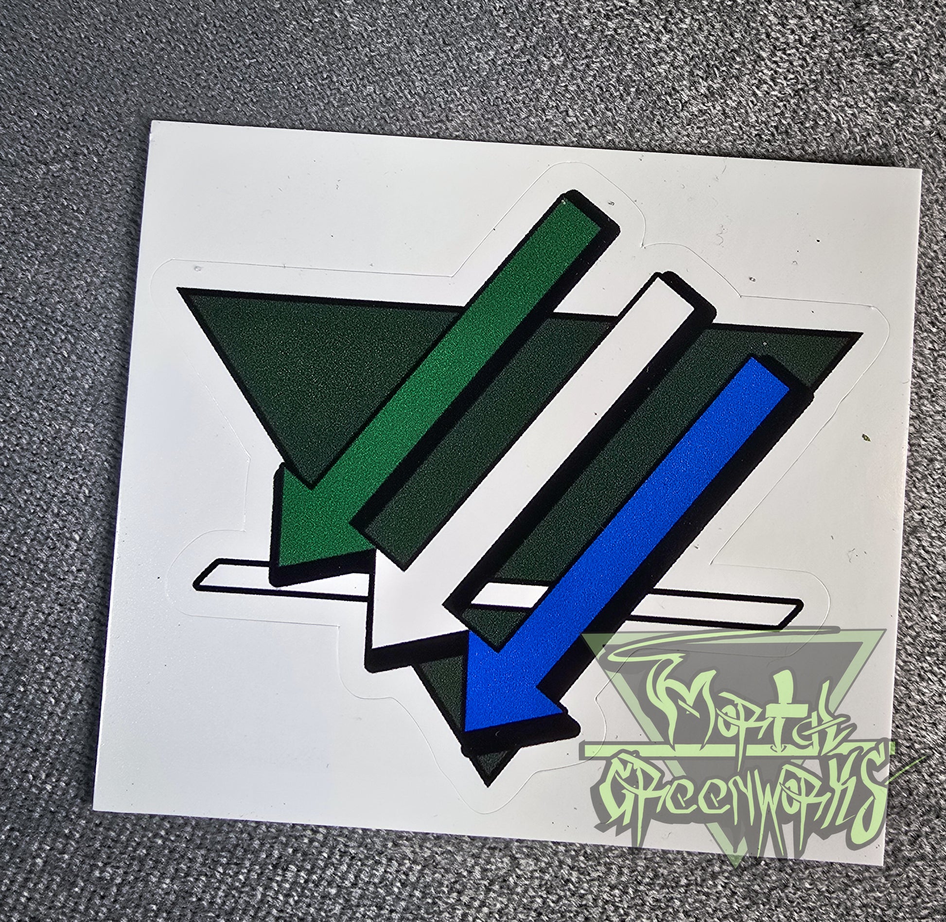 Photograph of a forest green 3x3" triangular sticker with three arrows and a white line overlaying. The arrows are from top to bottom green, white and blue.