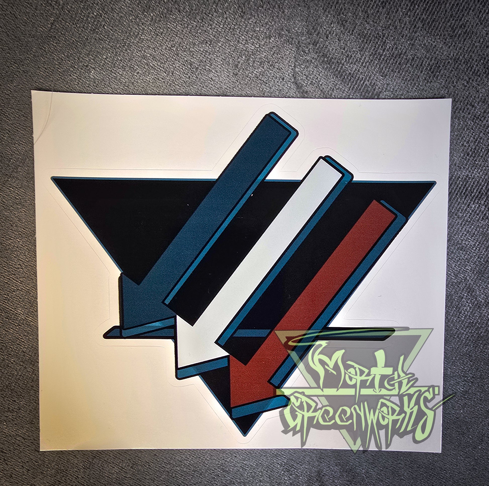 A 3x3" sticker is placed on a dark tablecloth. The sticker design consists of three turquoise, white, and brown downward arrows pointing to the left. Laid out on top of a black triangle turned upside down with a bold dark blue line in the bottom third.
