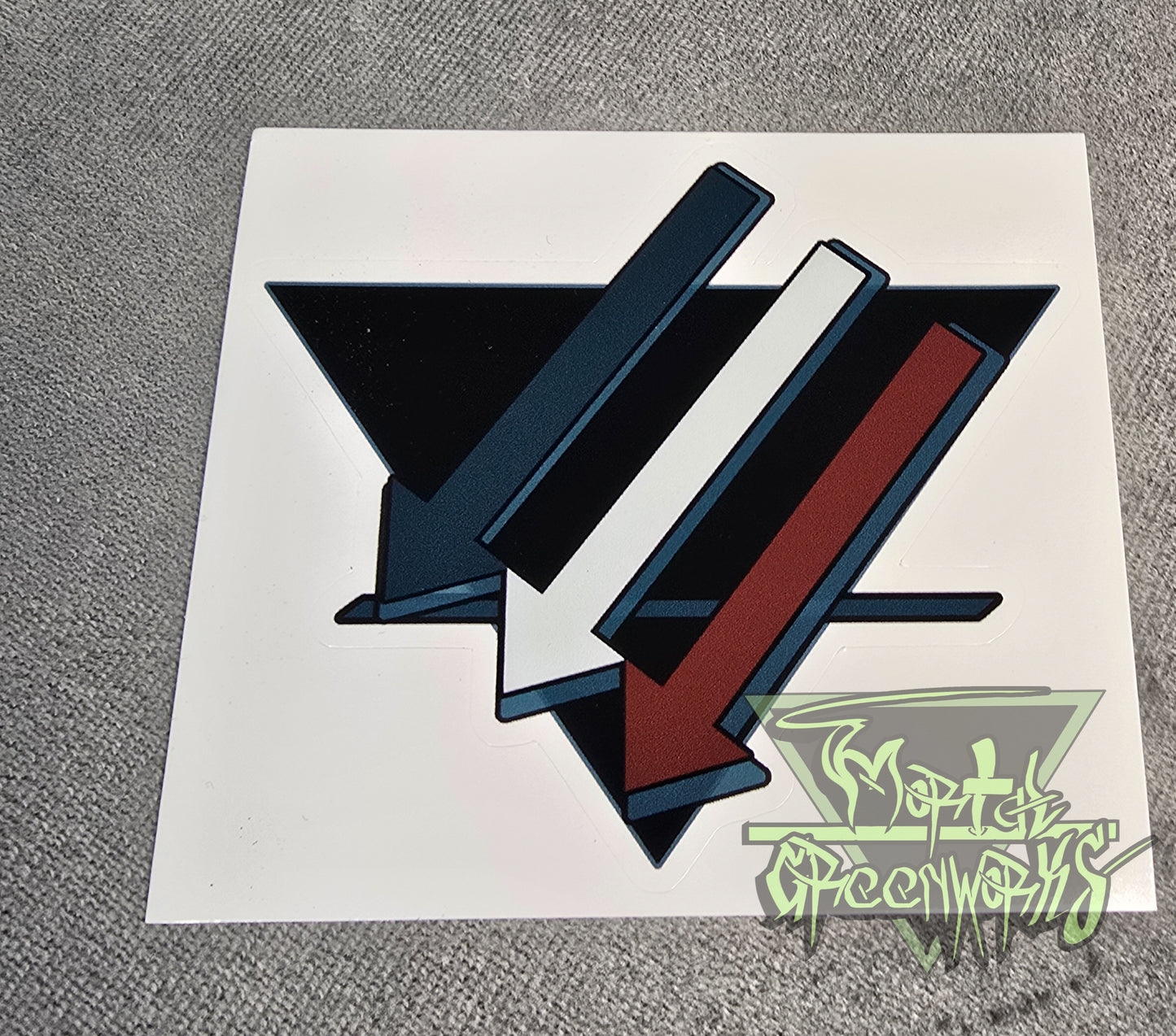 A 4x4" sticker is placed on a dark tablecloth. The sticker design consists of three turquoise, white, and brown downward arrows pointing to the left. Laid out on top of a black triangle turned upside down with a bold dark blue line in the bottom third.