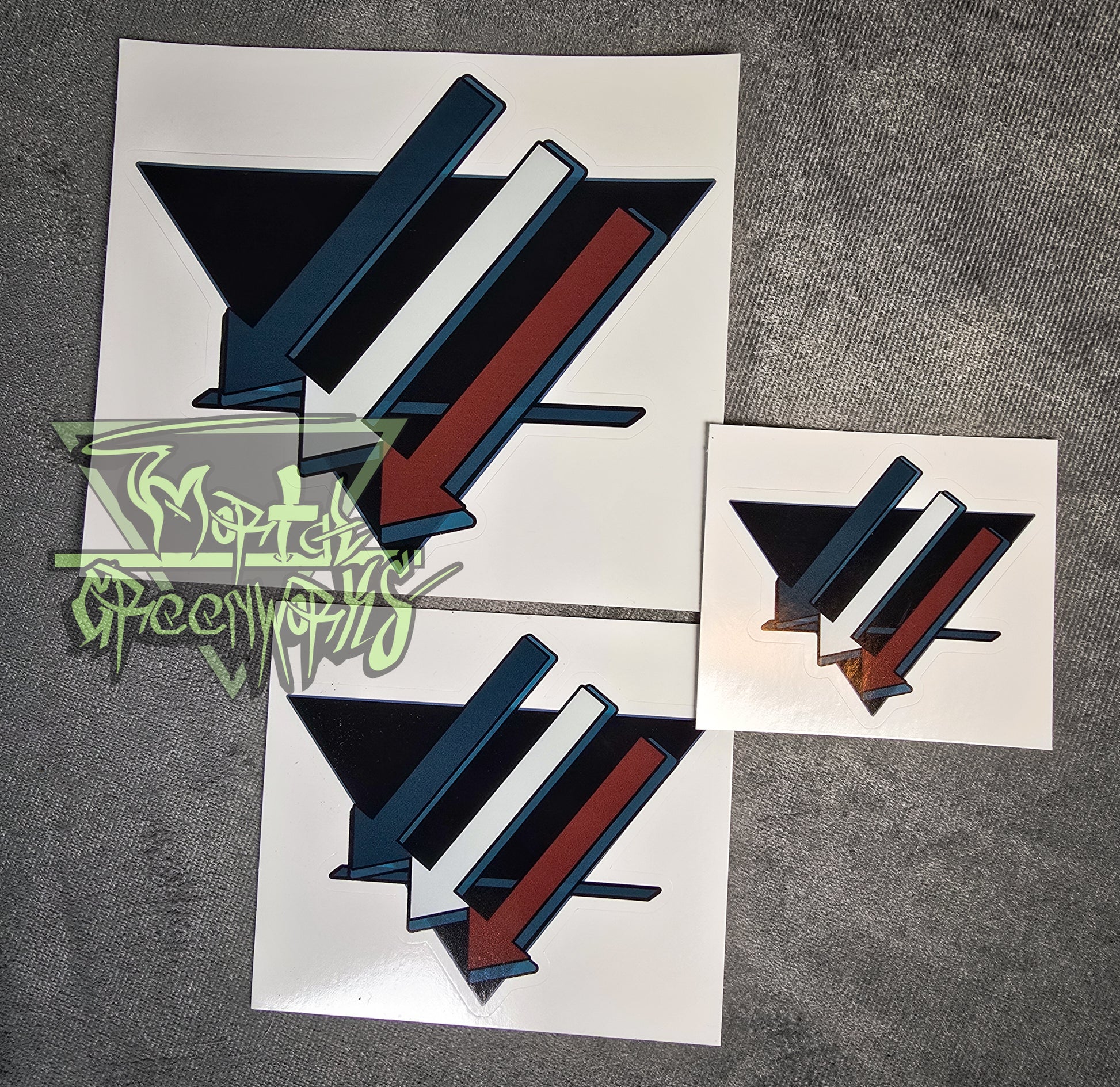 A large, medium and a small sticker are placed on a dark tablecloth. The sticker design consists of three turquoise, white, and brown downward arrows pointing to the left. Laid out on top of a black triangle turned upside down with a bold dark blue line in the bottom third. The Mortal Greenworks logo is in the bottom right hand side of the photograph.