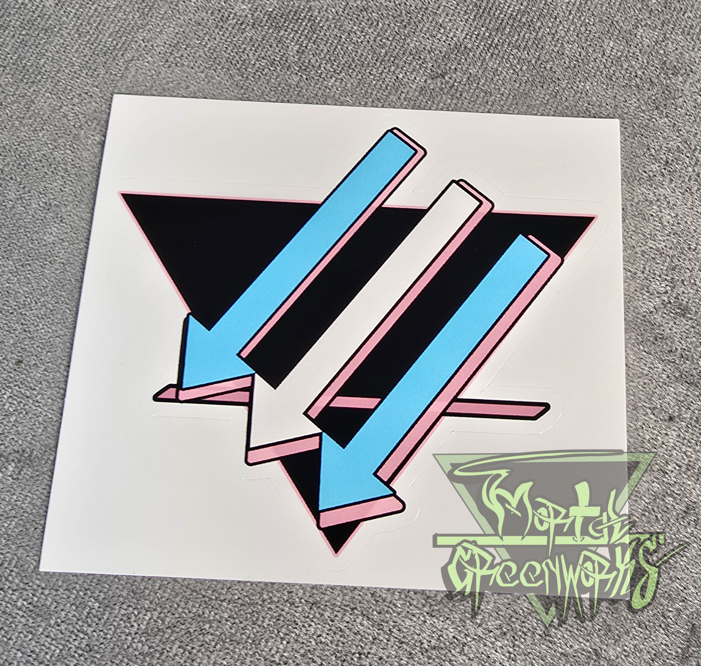 A close up photograph of a  3x3 sticker placed on a light grey tablecloth. The design is a black upside down triangle with a pink line in the bottom third with light blue and white arrows pointing left and downward. The Mortal Greenworks Watermark is in the bottom right hand corner of the image.