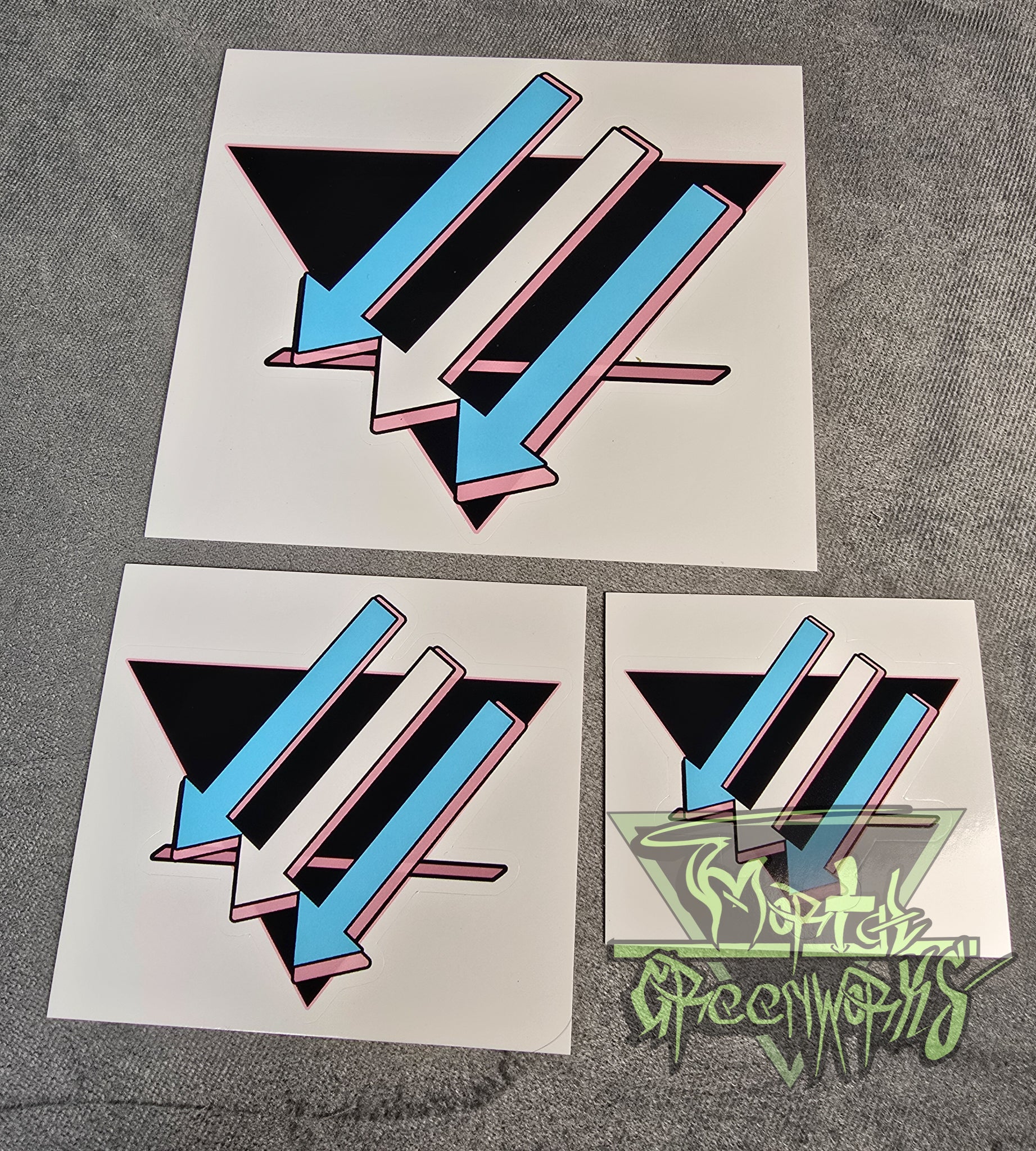 A close-up photograph of a  3x3", 4x4", and a 6x6" sticker placed on a light grey tablecloth. The design is a black upside down triangle with a pink line in the bottom third with light blue and white arrows pointing left and downward. The Mortal Greenworks Watermark is in the bottom right hand corner of the image.