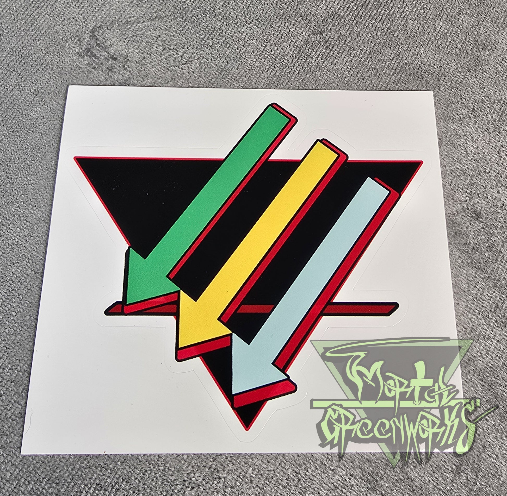Photo of a colorful geometric design on a 3x3 sticker sheet with a brand logo in the corner of the photo. Three arrows, top to bottom: light green, gold, and turquoise, are paired against a black upside-down triangle and a red line in the bottom third. 
