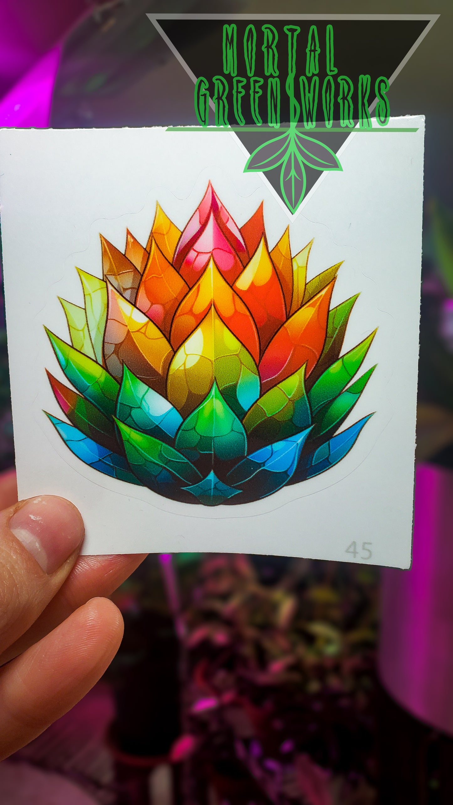 A sticker of a stylized agave plant with leaves in a gradient of cosmic colors, held by a hand with the 'Mortal Greenworks' logo above.