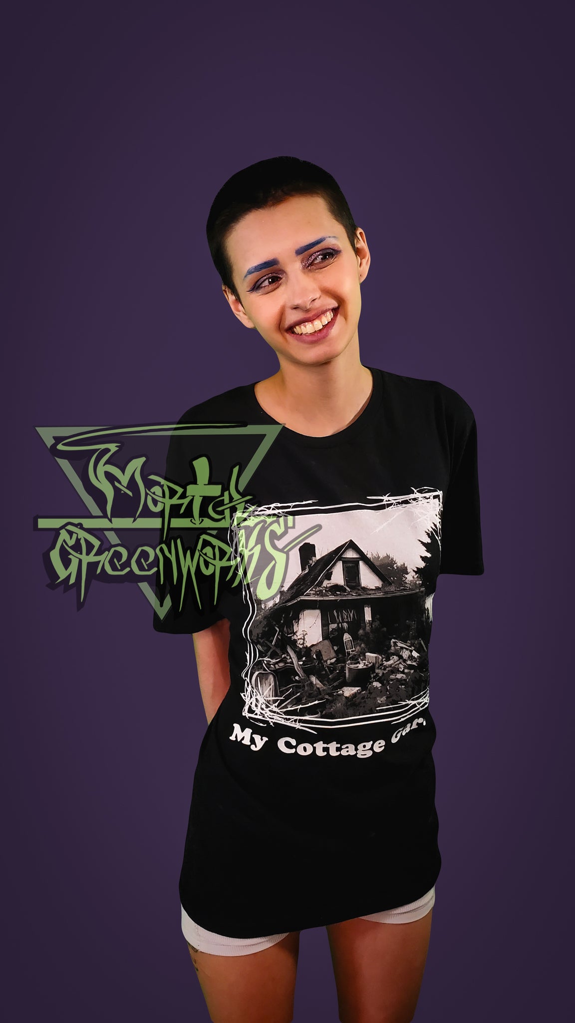 A smiling person wearing a black t-shirt with a cottage graphic design on a purple background.