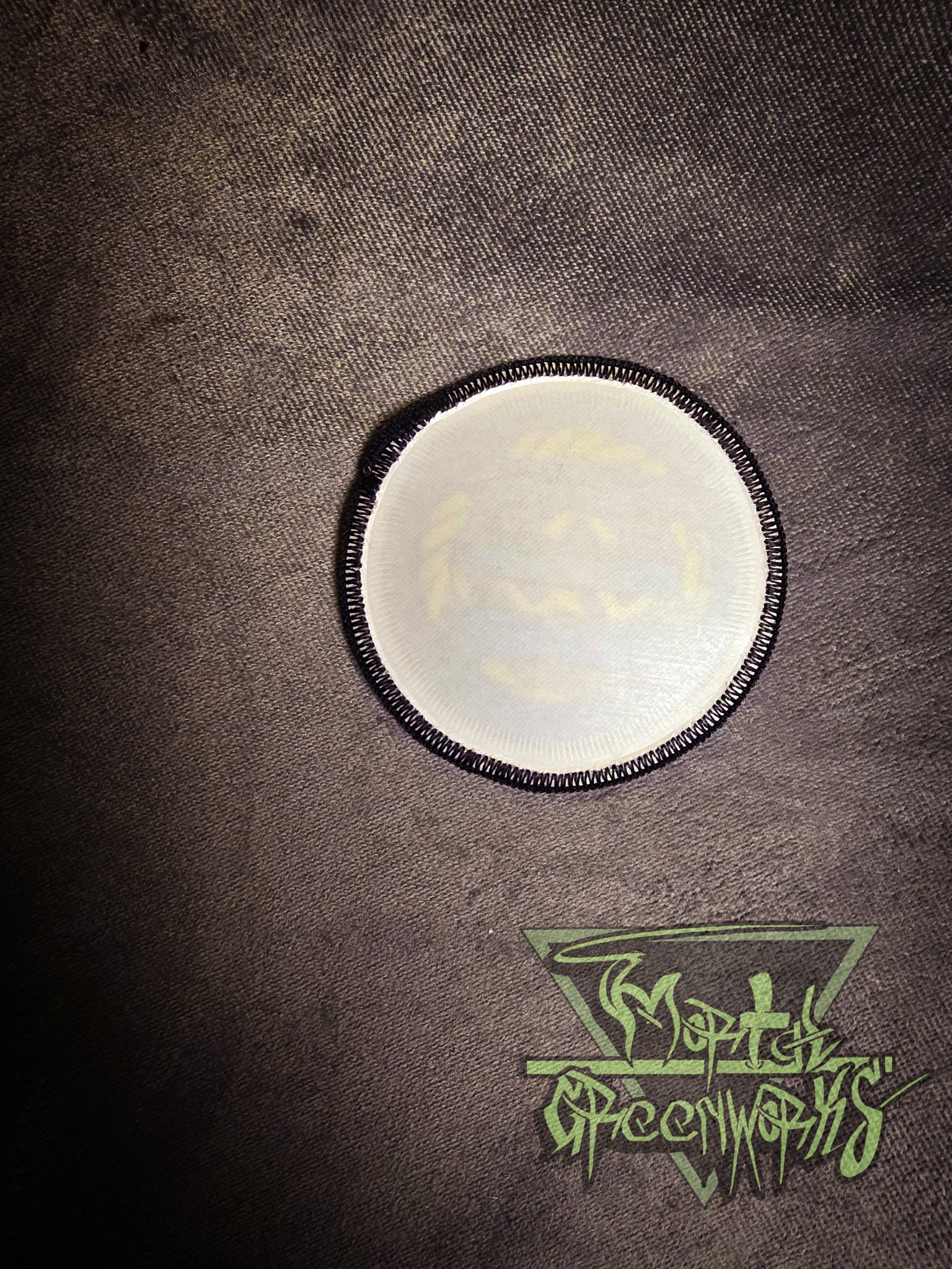 The backside of a round white iron-on patch with a black border on a textured brown surface, featuring a logo at the bottom.