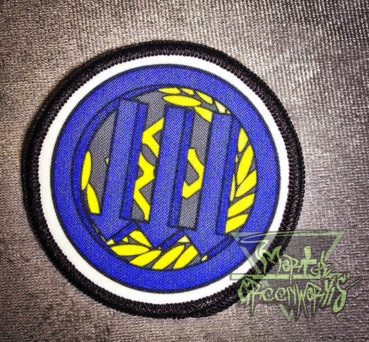 Circular patch with black and white rim, with three arrows pointing down over the golden PB logo and laurel wreath. Set on a grey tablecloth with a brand logo in the corner of the photo
