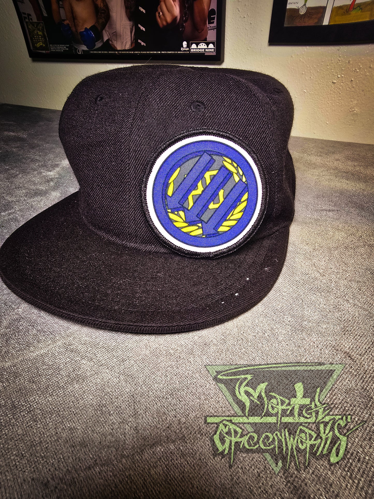 Photo of a black baseball cap rseting on a table with punk band posters in the background. Resting on the hat is a circular patch with black and white rim, with three arrows pointing down over the golden PB logo and laurel wreath.