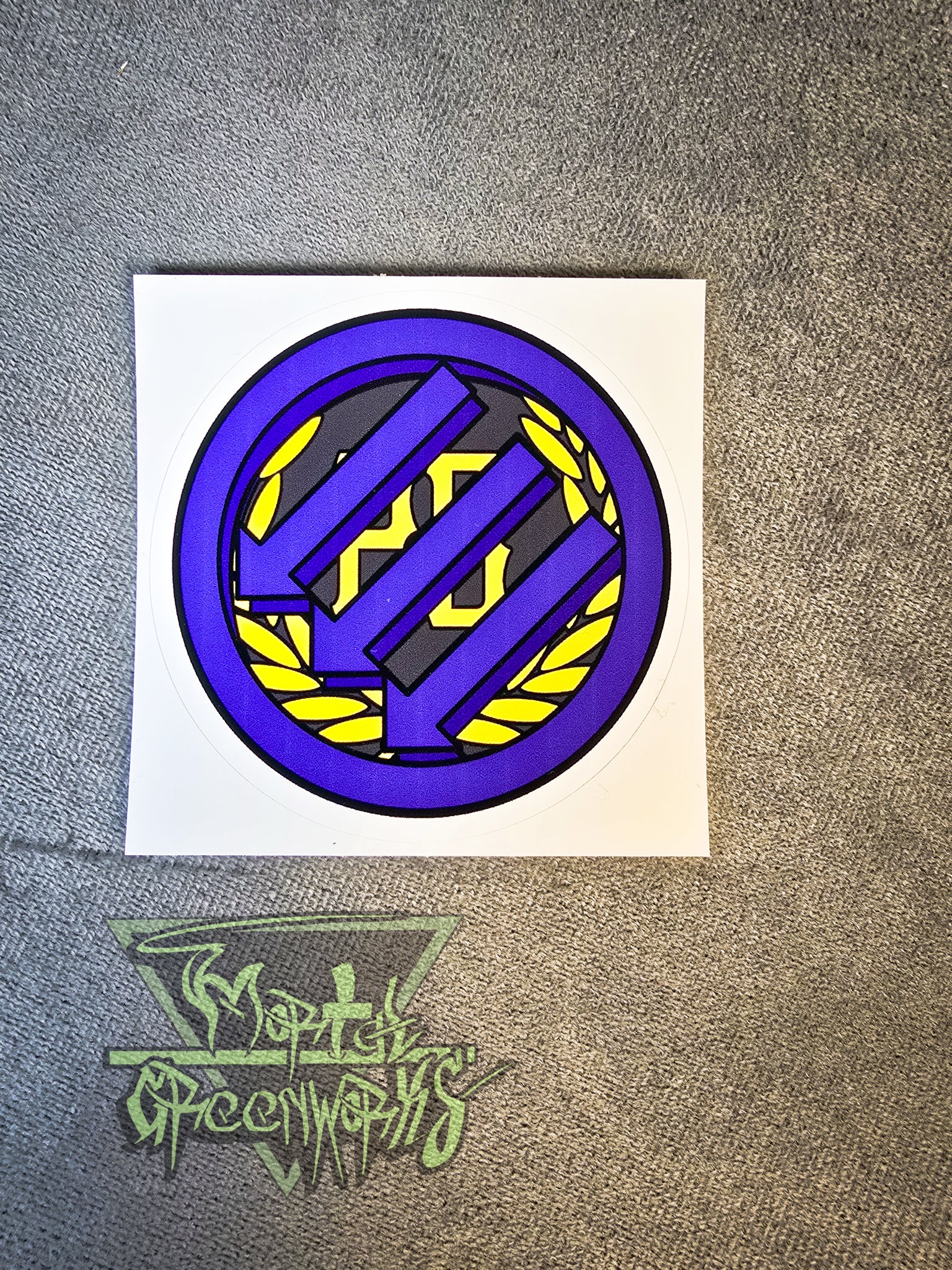 A 3"x3" sticker sits on a grey tablecloth, showing the Iron Front logo painted purple atop a gold Proud Boy logo. The Mortal Greenworks logo is in the bottom left corner.