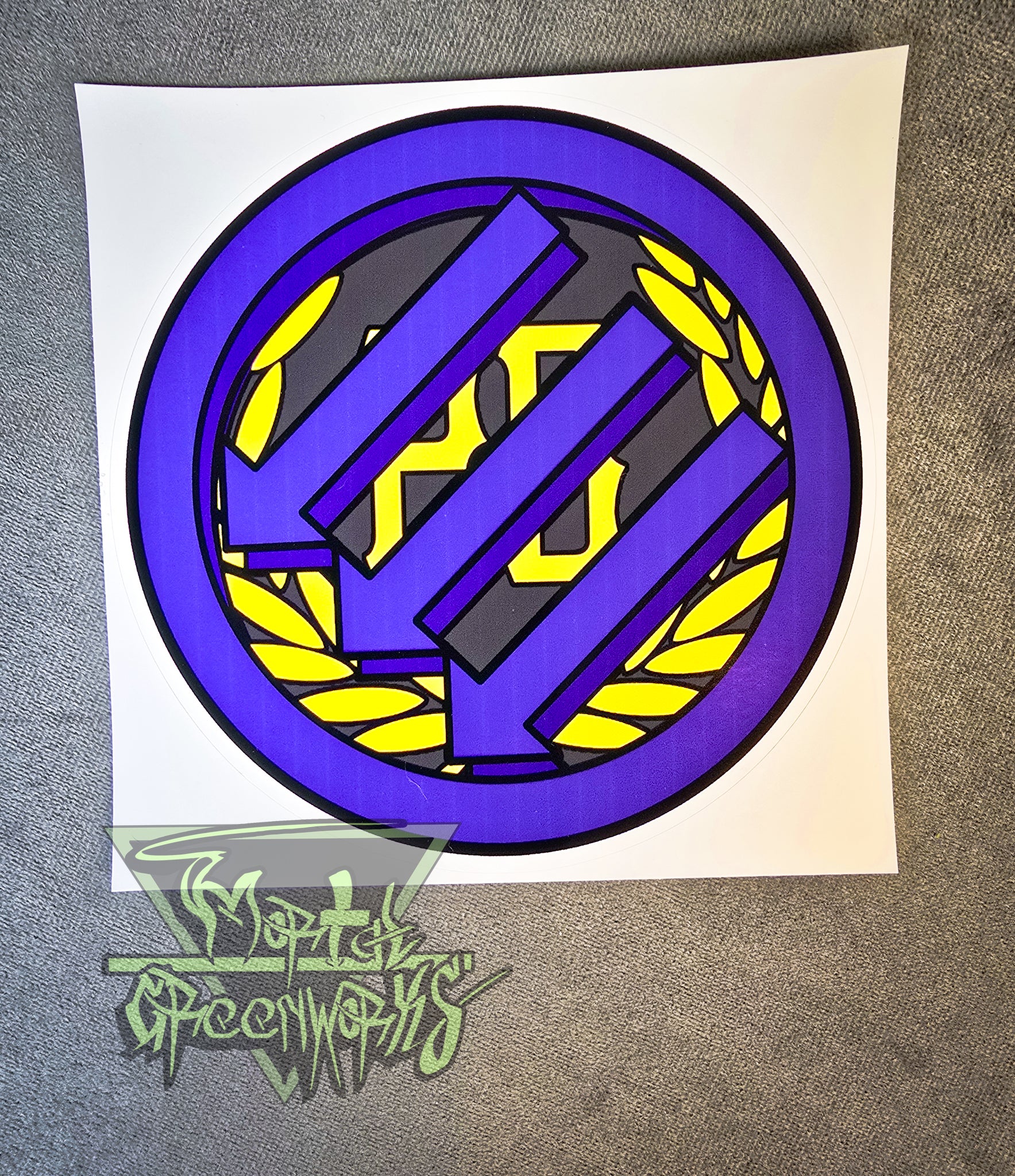 A 4"x4" sticker sits on a grey tablecloth, showing the Iron Front logo painted purple atop a gold Proud Boy logo. The Mortal Greenworks logo is in the bottom left corner.