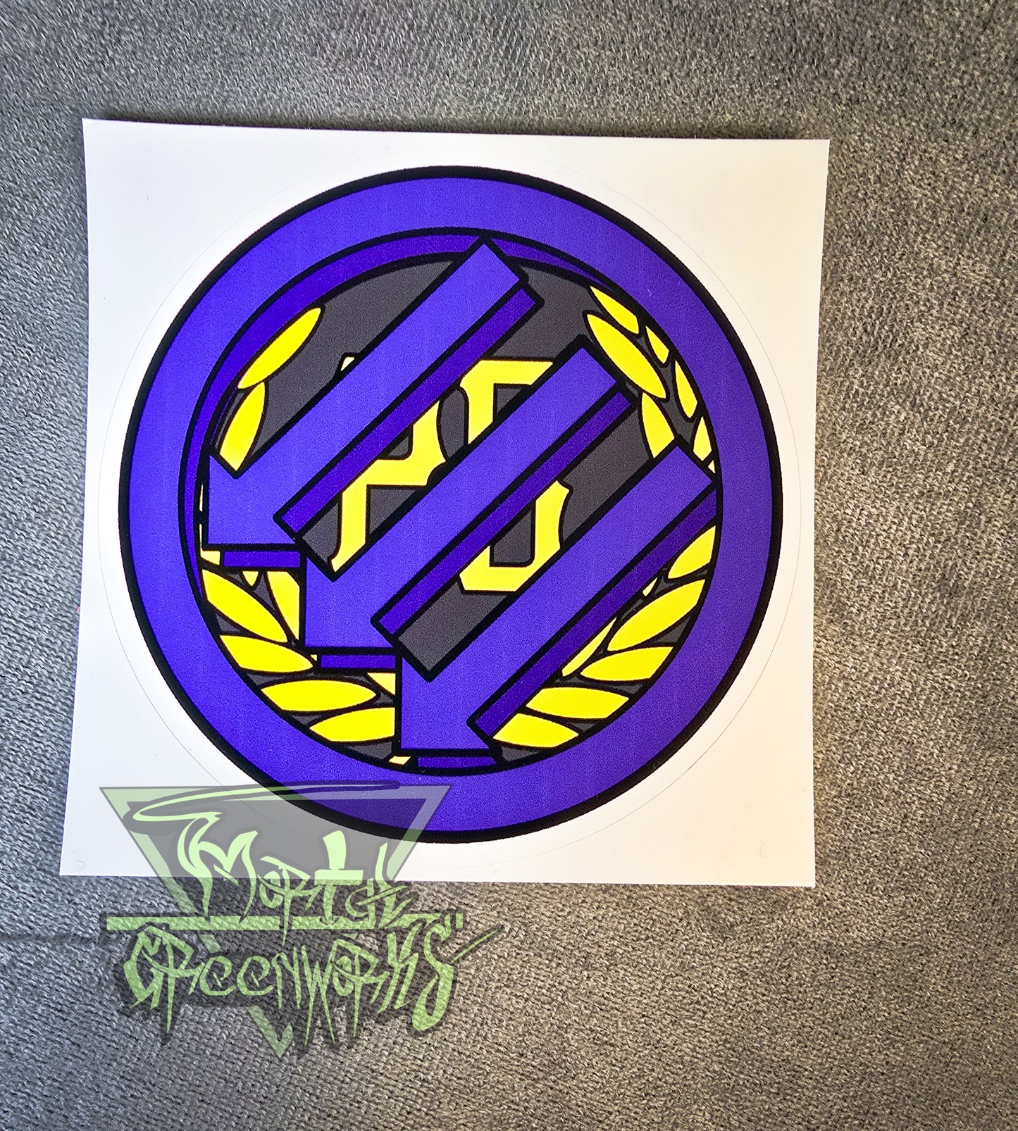 A 6"x6" sticker sits on a grey tablecloth, showing the Iron Front logo painted purple atop a gold Proud Boy logo. The Mortal Greenworks logo is in the bottom left corner.