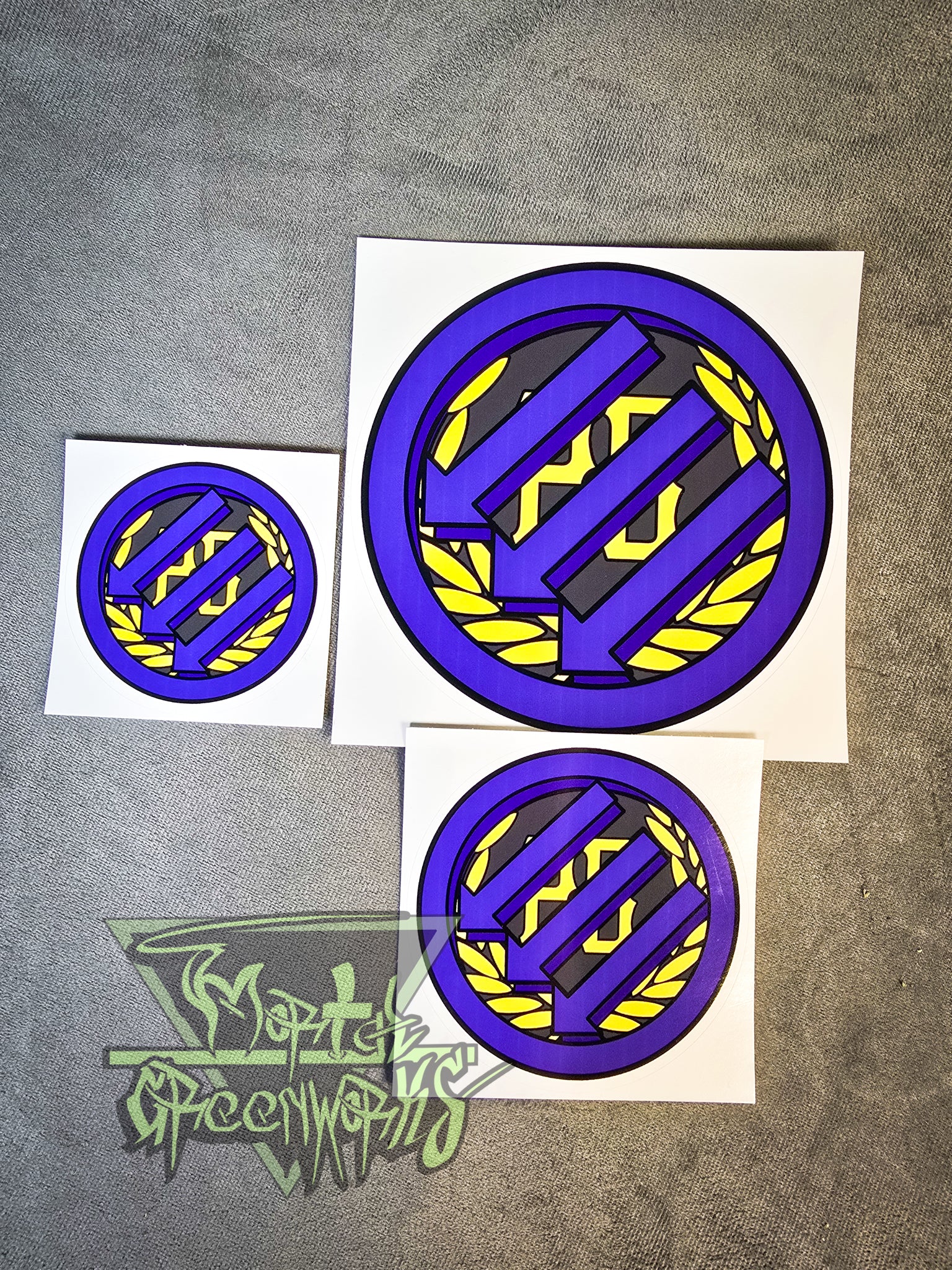 Three stickers sit on a grey tablecloth, showing the Iron Front logo painted purple atop a gold Proud Boy logo. The Mortal Greenworks logo is in the bottom left corner. All three sizes, from 3x3", 4x4", and 6x6", are displayed side by side.